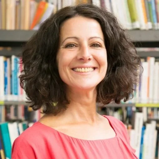 Mujer sonriendo con cabello rizado, vestida con blusa rosa, en una biblioteca.