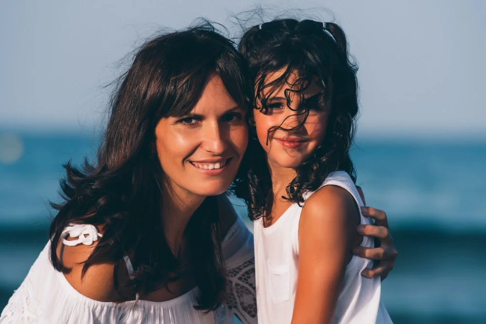 Mujer y niña abrazándose en la playa con el mar de fondo, ambas sonriendo.