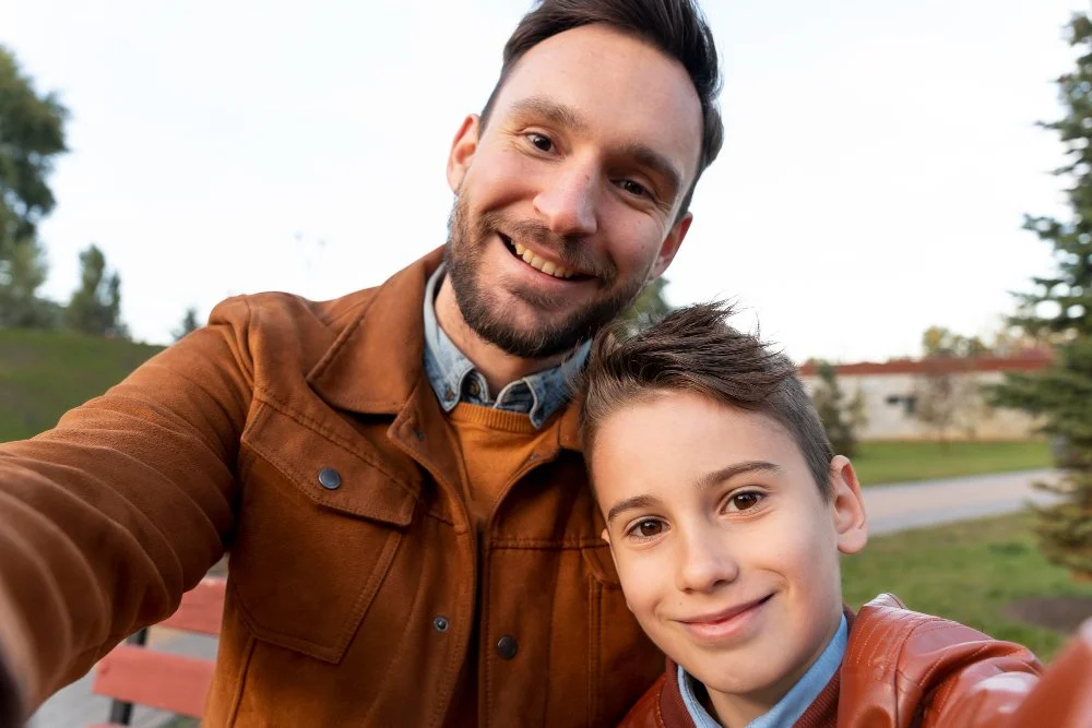Un hombre y un niño sonriendo, tomándose una selfie en un parque al aire libre, con árboles y césped de fondo.