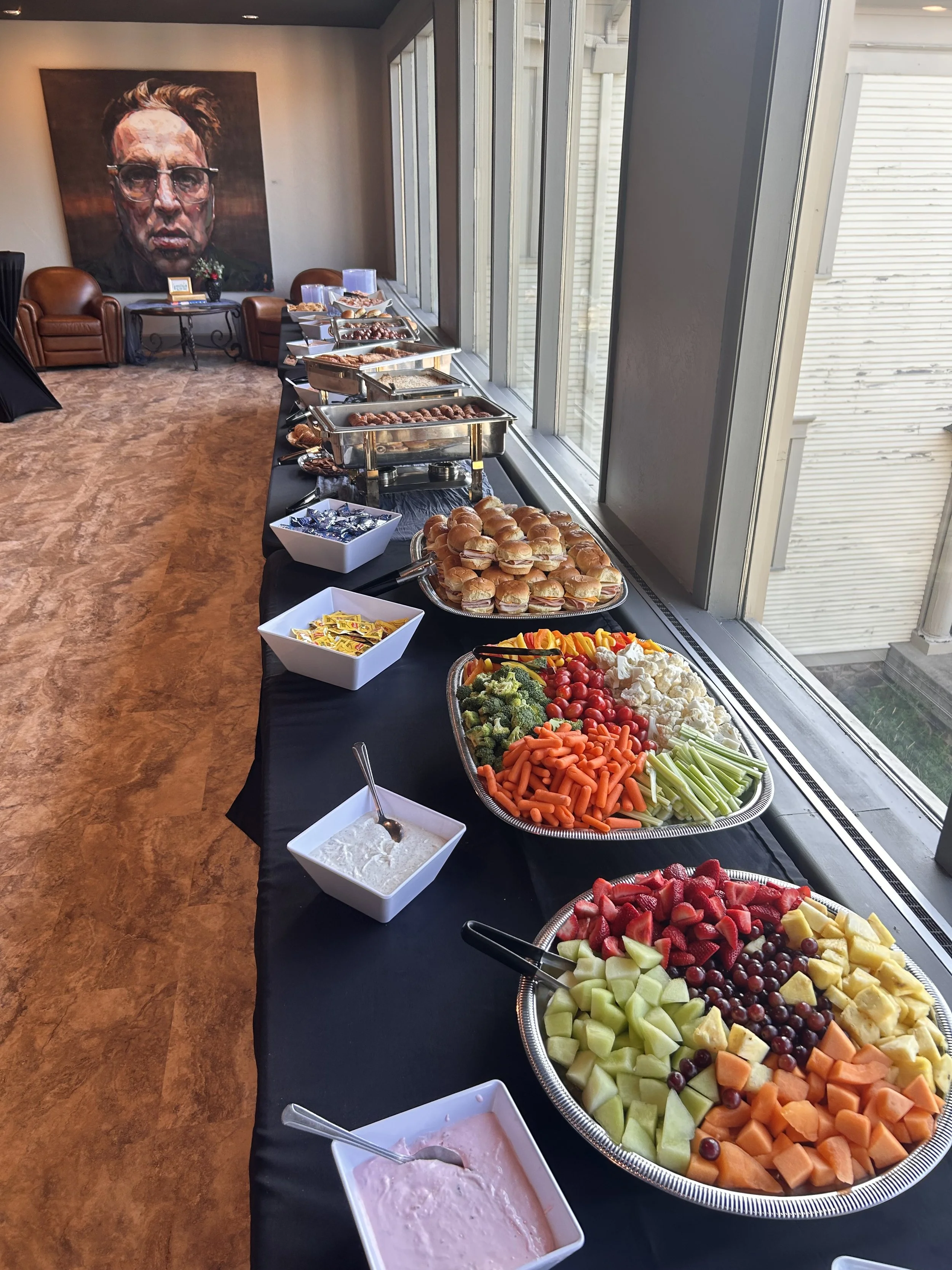 Buffet table with an assortment of fresh fruits, vegetables, and dips, set next to a large window in a room with a portrait painting on the wall.