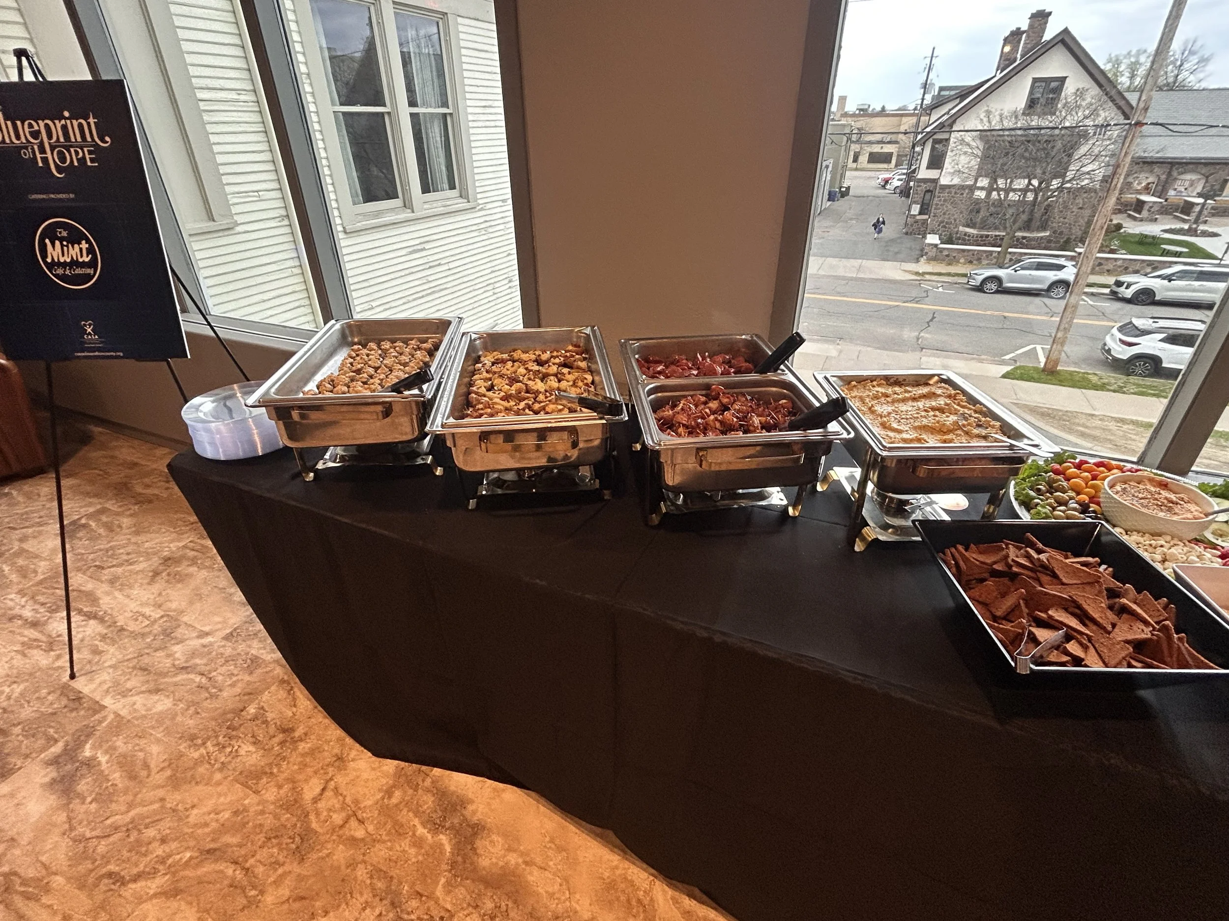 Buffet table with trays of food, including meats, pasta, and side dishes, set near a large window overlooking a street with parked cars and buildings.