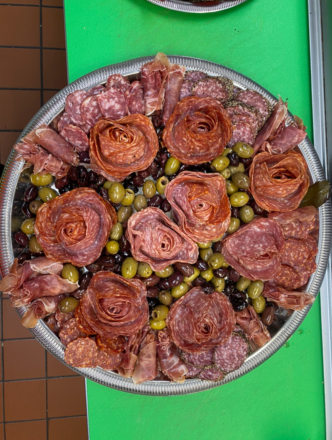 A large round serving tray with assorted slices of cured meats, including salami, prosciutto, and pepperoni, garnished with green and black olives, placed on a green cutting board.