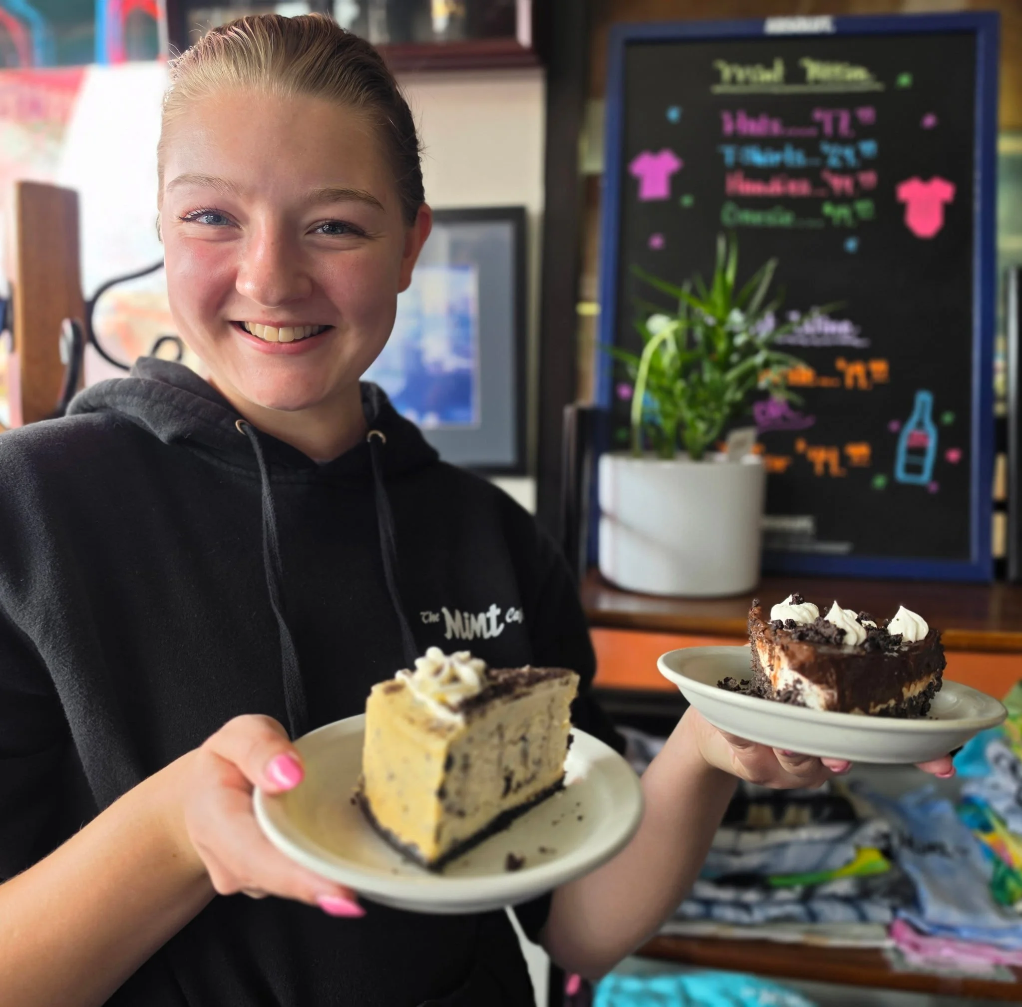 A young girl with blonde hair in a ponytail holding up two small cups of dessert in a store or cafe. One cup contains a layered cake with chocolate and cream, and the other has a yellowish dessert with dark crumbs and dollops of whipped cream.