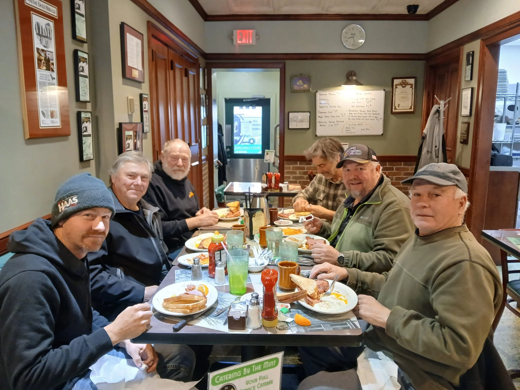 Group of six men sitting at a restaurant table enjoying breakfast with plates of toast, eggs, and bacon, surrounded by condiments, cups, and glasses, inside a cozy, decorated dining area.