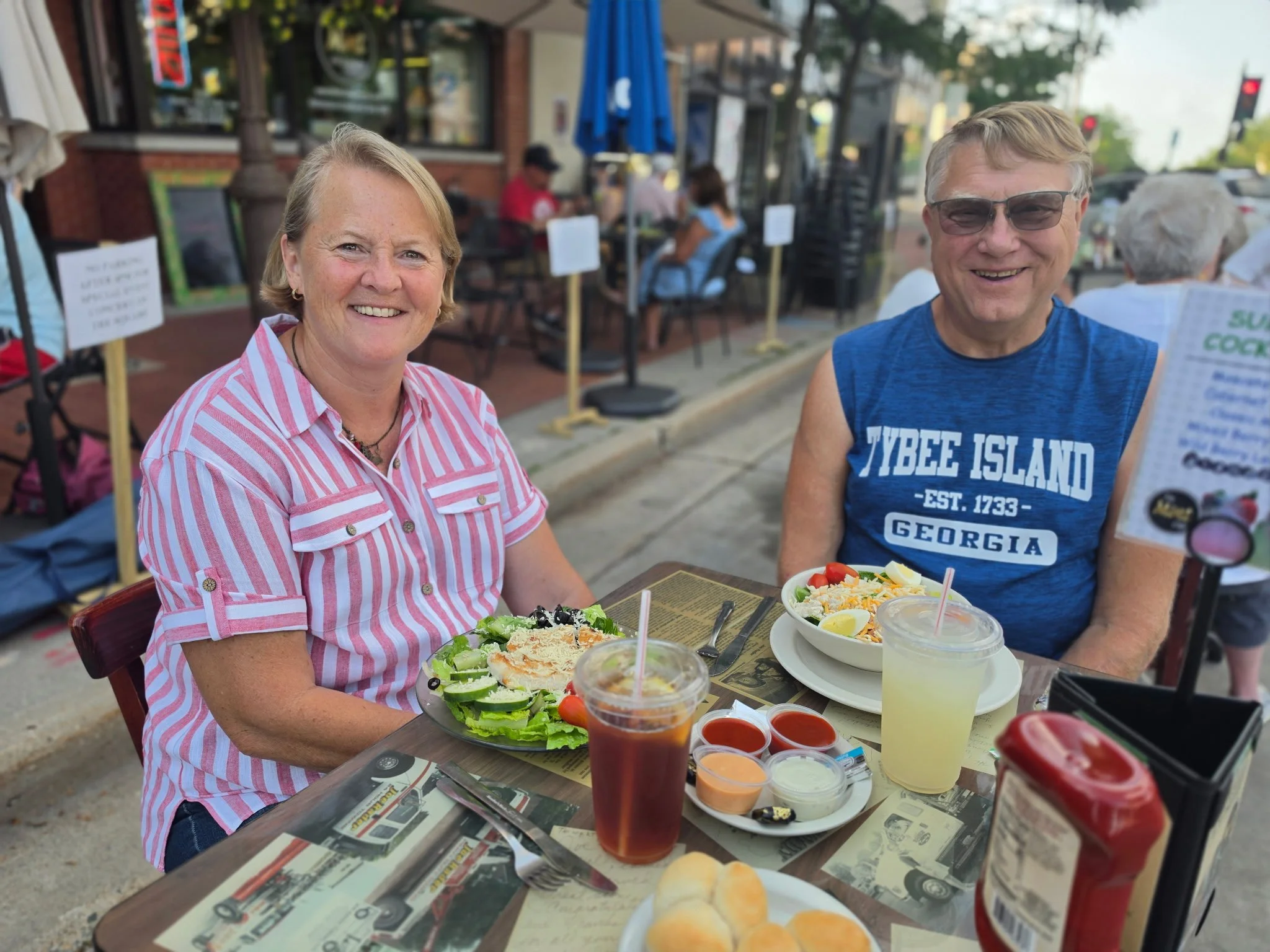 A smiling woman in a pink striped shirt and a smiling man in a blue sleeveless shirt with glasses sit at an outdoor restaurant table with salads and drinks. There are other diners and a building with large windows in the background.