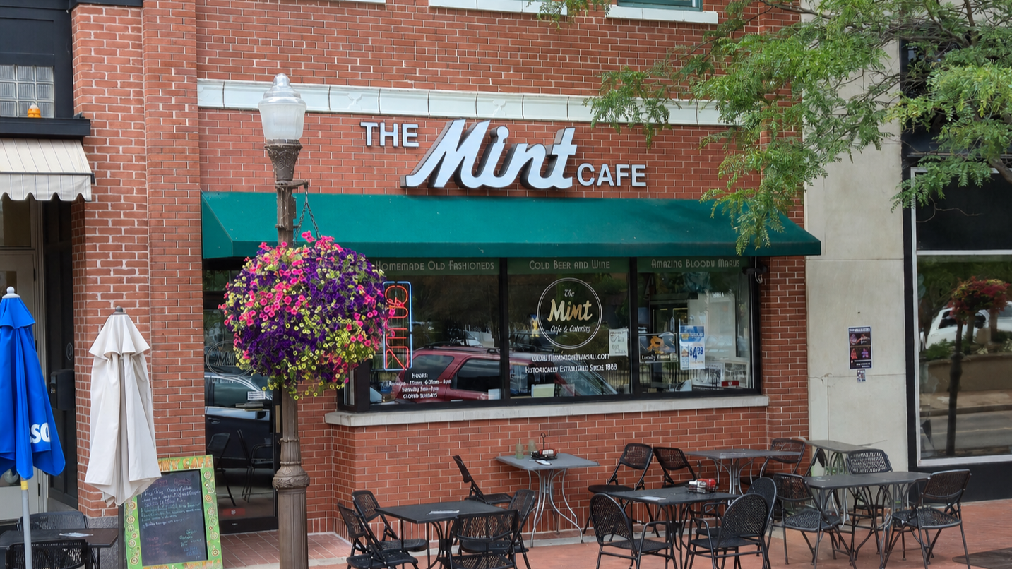 Exterior of The Mint Cafe with outdoor seating, a hanging flower basket, and colorful signage on a brick building.