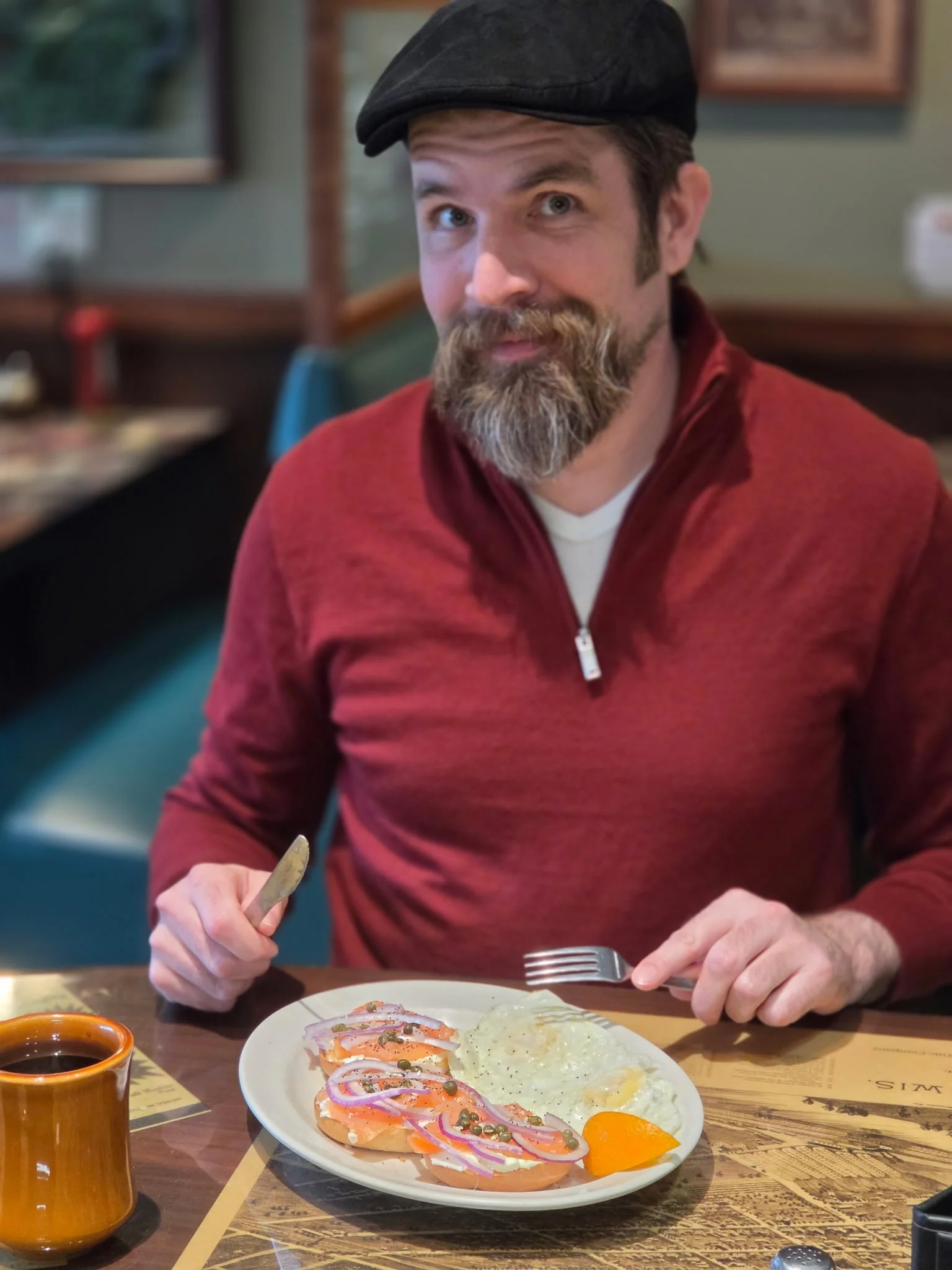 A man with a beard and a black cap is sitting at a table in a restaurant, preparing to eat a plate of food that includes eggs, sliced tomatoes, onion, and possibly fish or other protein, with a small bowl of sauce and a cup of coffee nearby.