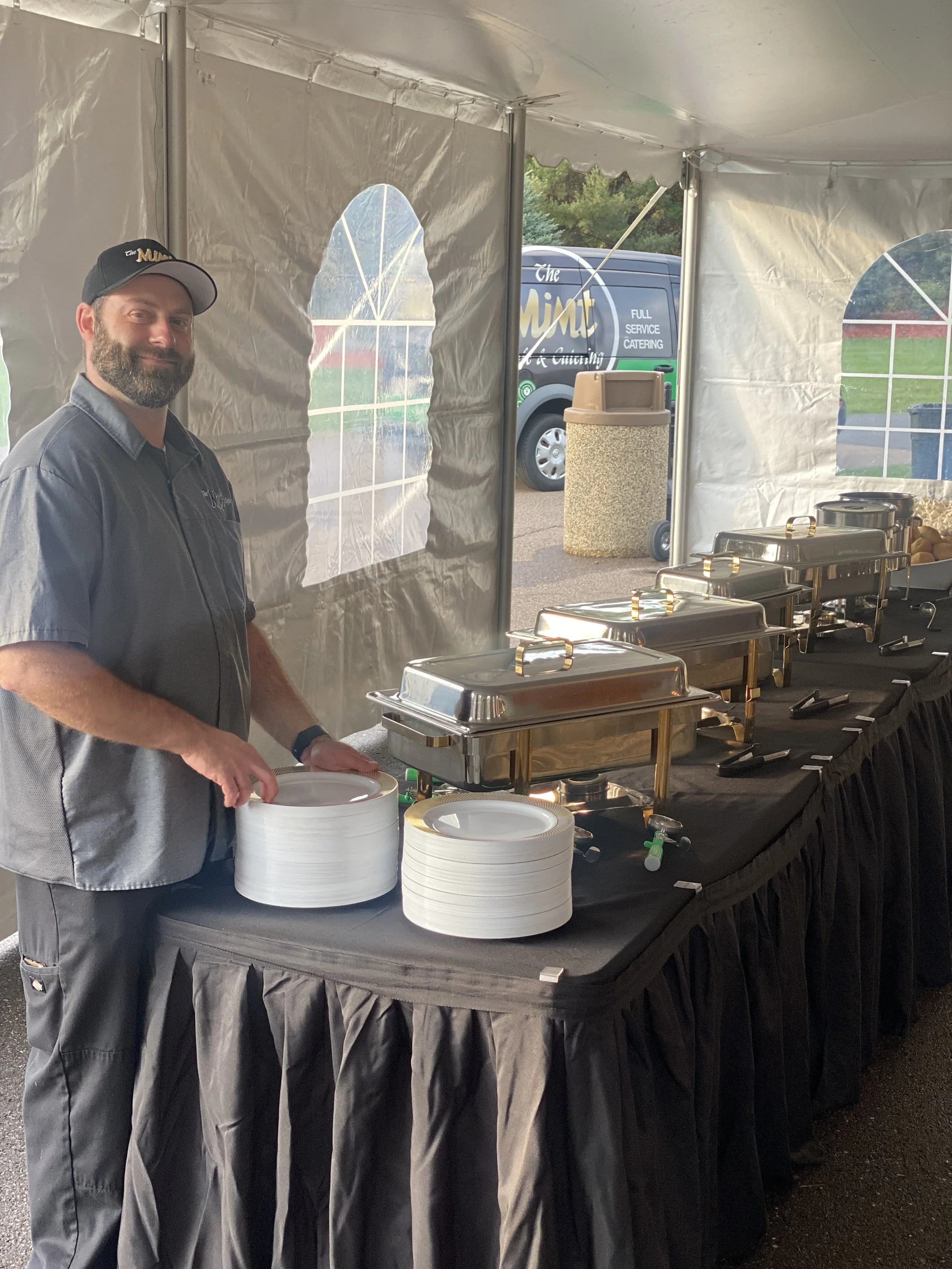 Man with a beard wearing a black cap and gray shirt stands at a buffet table with chafing dishes inside a white event tent. The table has stacks of white plates and serving utensils. There is a catering van outside with logo and text visible, and a l