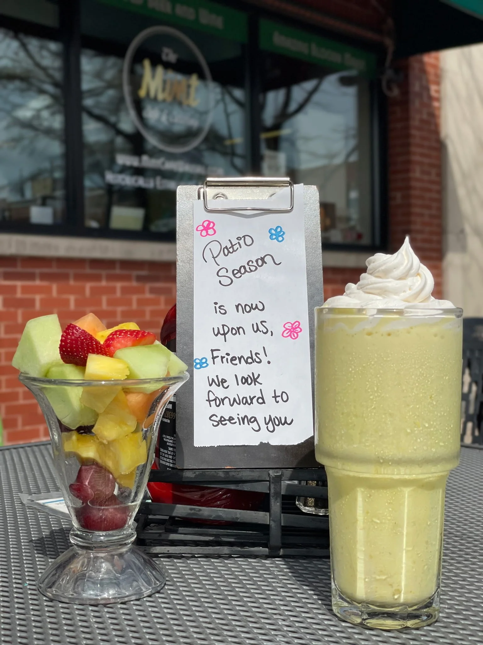 A clear glass bowl filled with various fresh cut fruits like strawberries, cantaloupe, honeydew, and pineapple, and a tall yellow smoothie or milkshake topped with whipped cream, placed on a gray metal surface outdoors. A handwritten sign on a clipbo