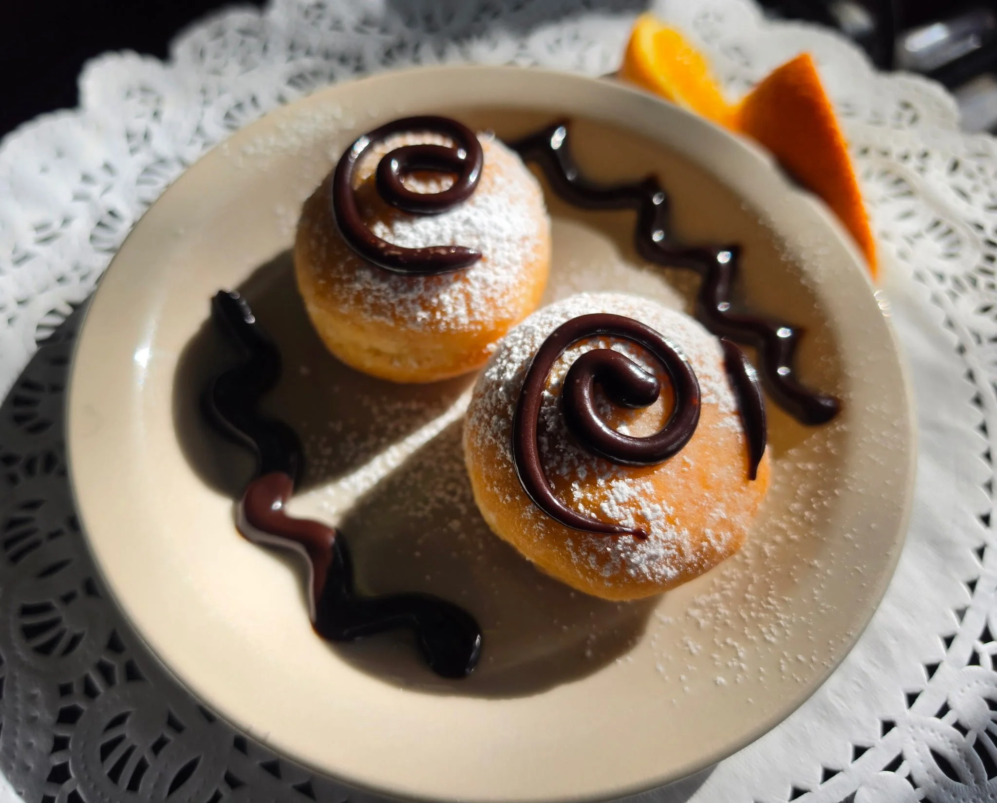 Plate with small round desserts dusted with powdered sugar, decorated with chocolate swirls, on a doily-lined surface with orange and lemon wedges in the background.