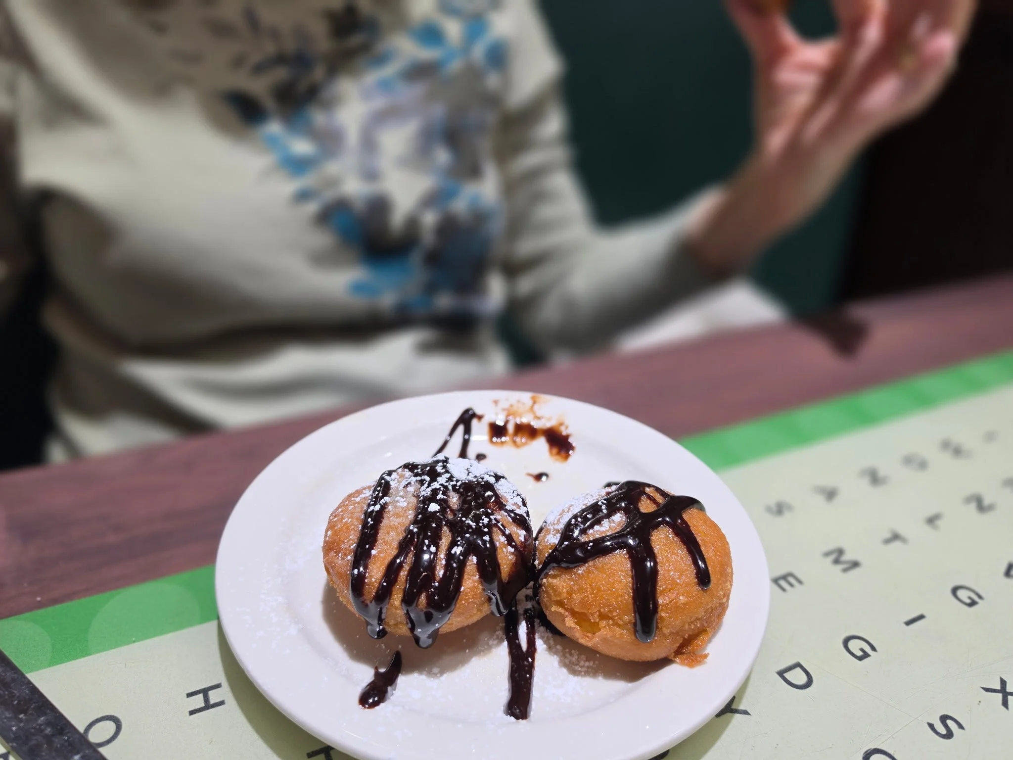 Two donuts with chocolate drizzle and powdered sugar on a white plate set on a table.