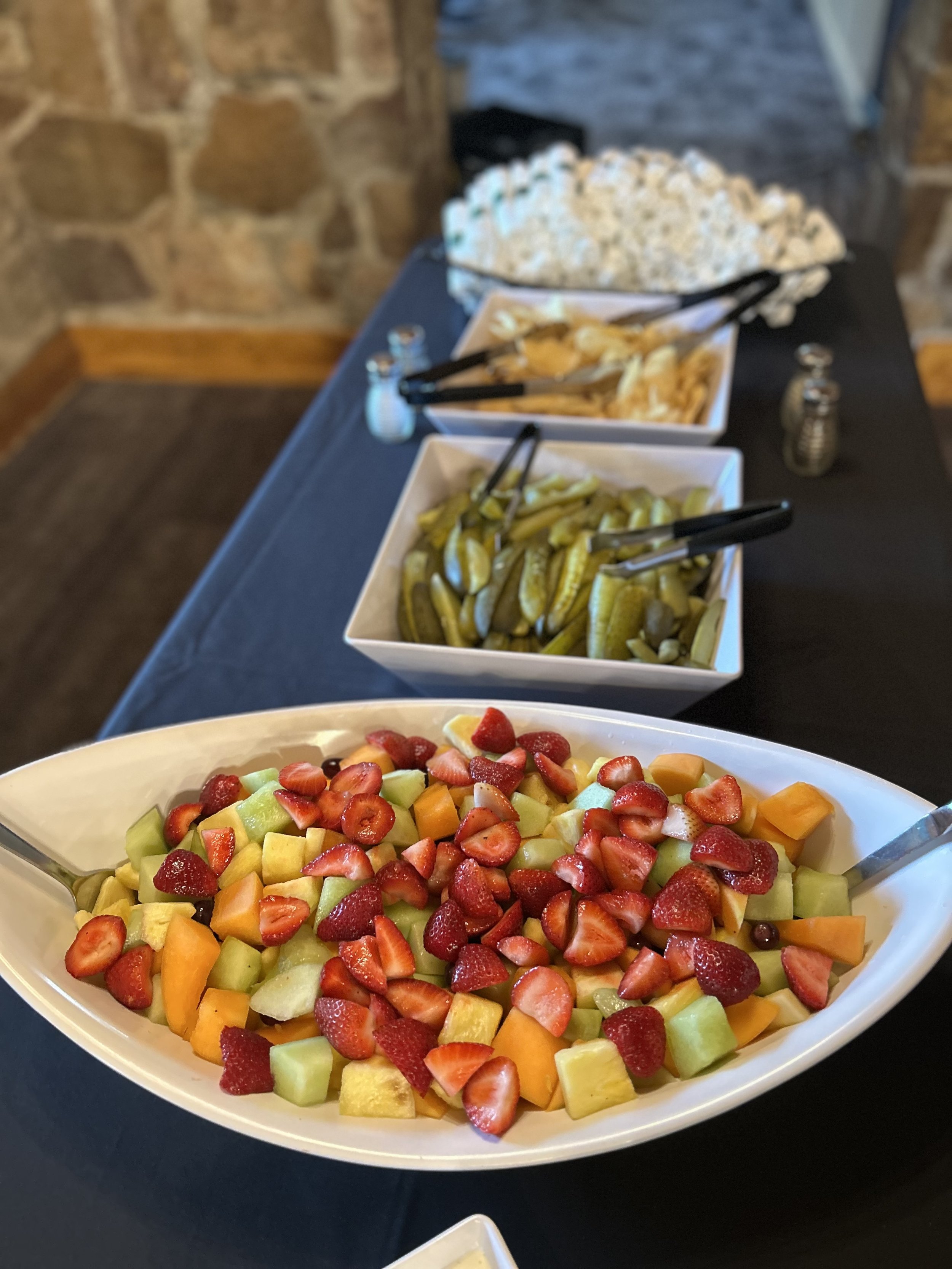 A large oval white bowl filled with chopped fresh fruit including strawberries, cantaloupe, honeydew, and pineapple, placed on a black table.