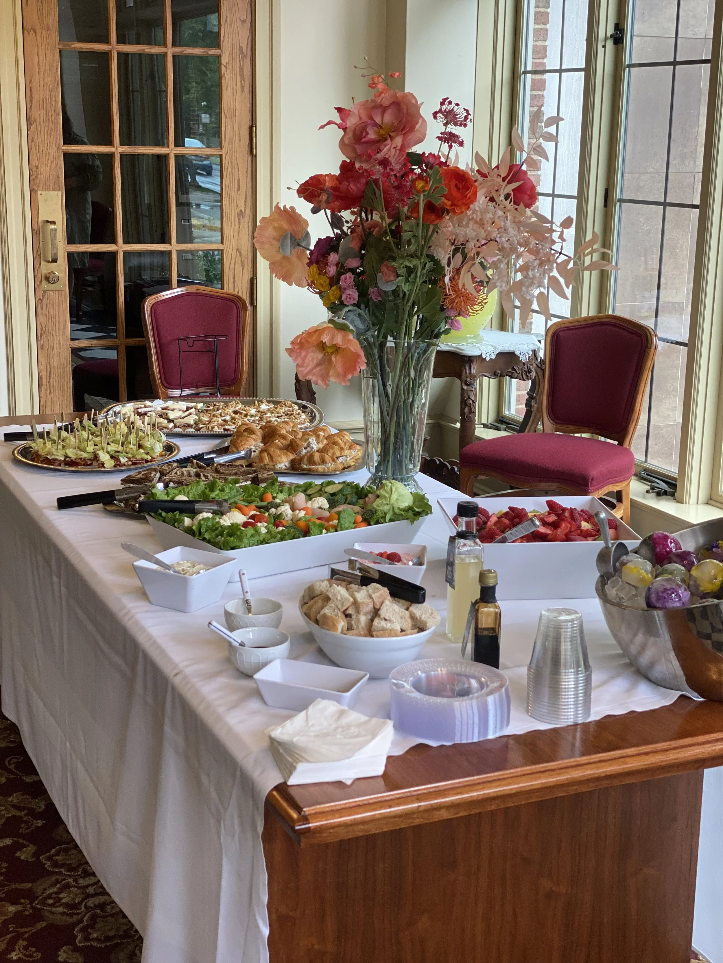 A dining table with a white tablecloth set for a meal, featuring a large glass vase of pink and red flowers as a centerpiece, surrounded by various dishes including salads, bread, strawberries, and condiments. There are two red upholstered chairs and