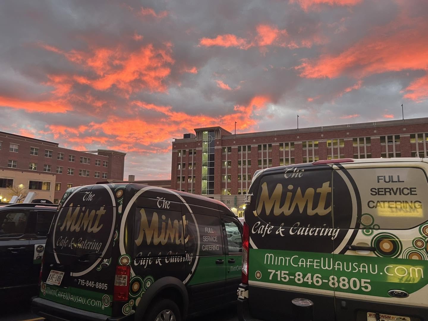 Two food trucks parked in front of a large brick building at sunset, with a colorful sky in the background.