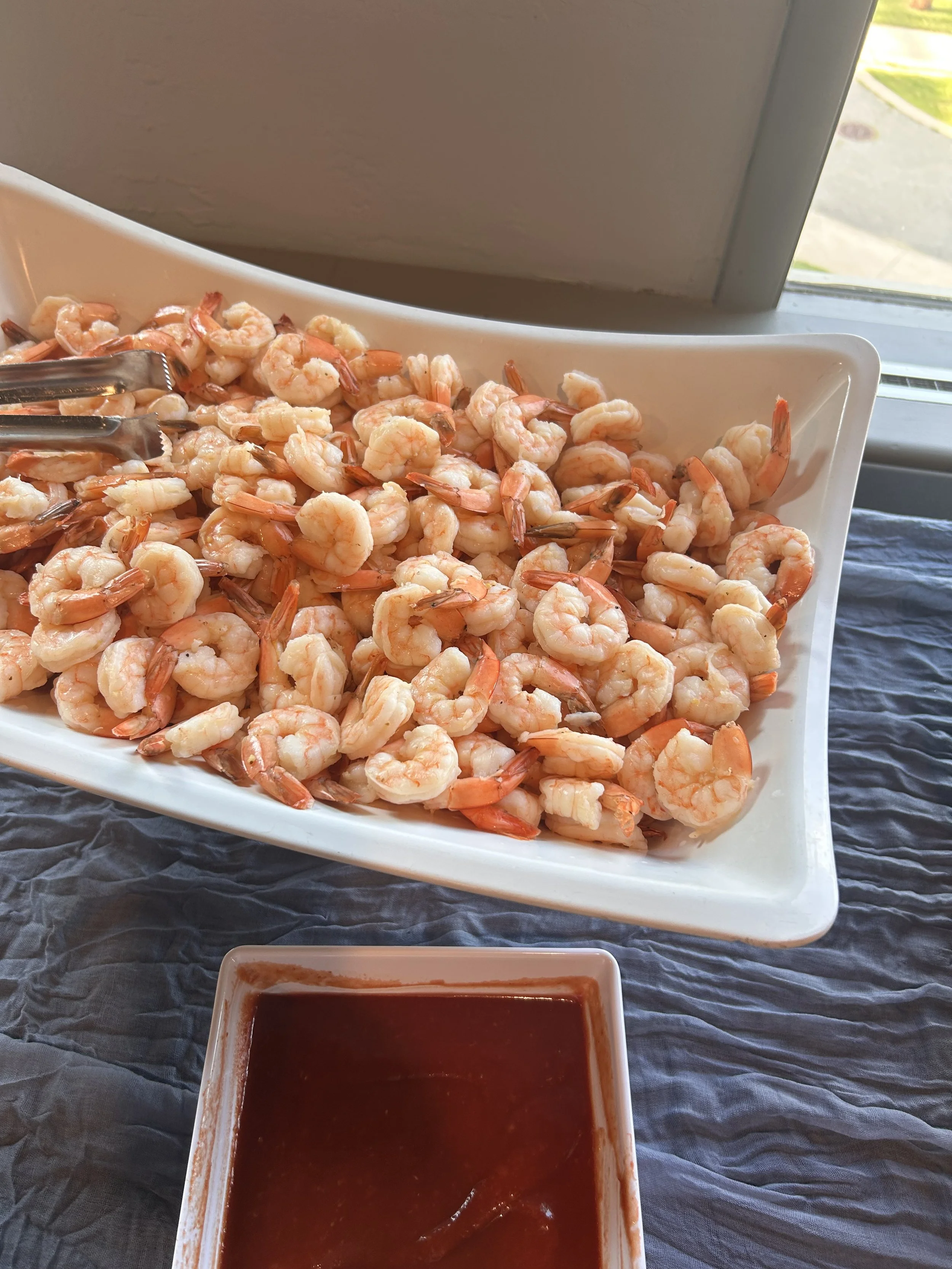 A white rectangular platter filled with cooked shrimp, placed on a table with a gray tablecloth near a window. In front of the platter, there is a small dish of dark dipping sauce.