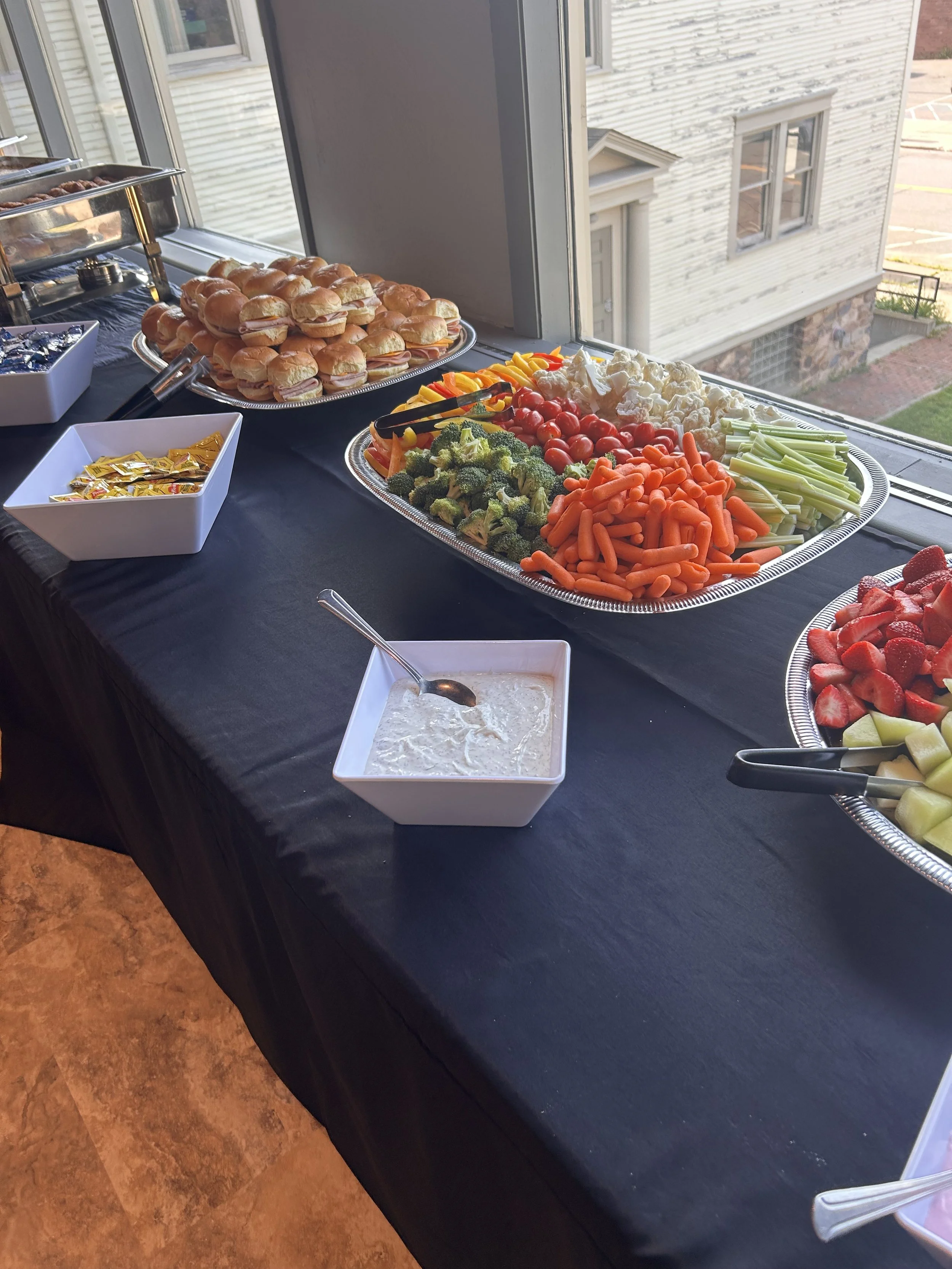 A buffet table with trays of vegetables, sandwiches, and condiments set by a large window, overlooking a residential building outside.