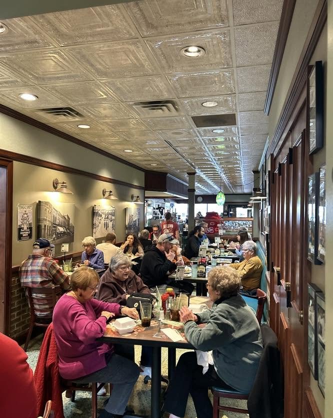 Interior of a busy restaurant with customers dining at tables, some eating and others conversing, with framed photographs on the wall and a green decorative hanging light in the background.
