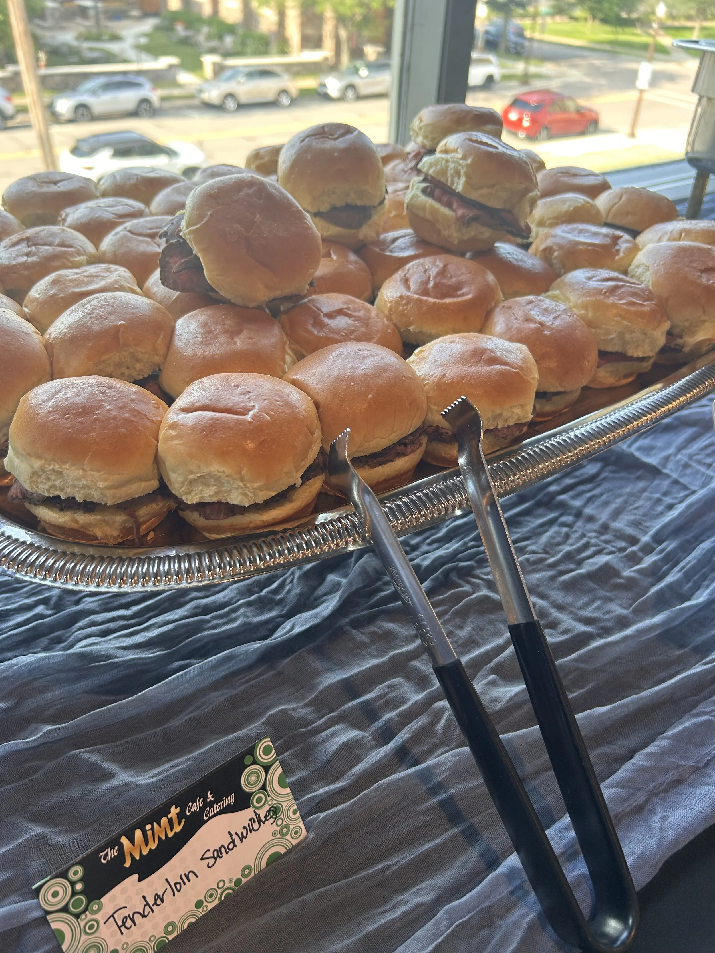 Close-up of a tray of small sandwich sliders and mini burgers on a silver platter with tongs, set on a table with a dark tablecloth near a window overlooking a parking lot and street.