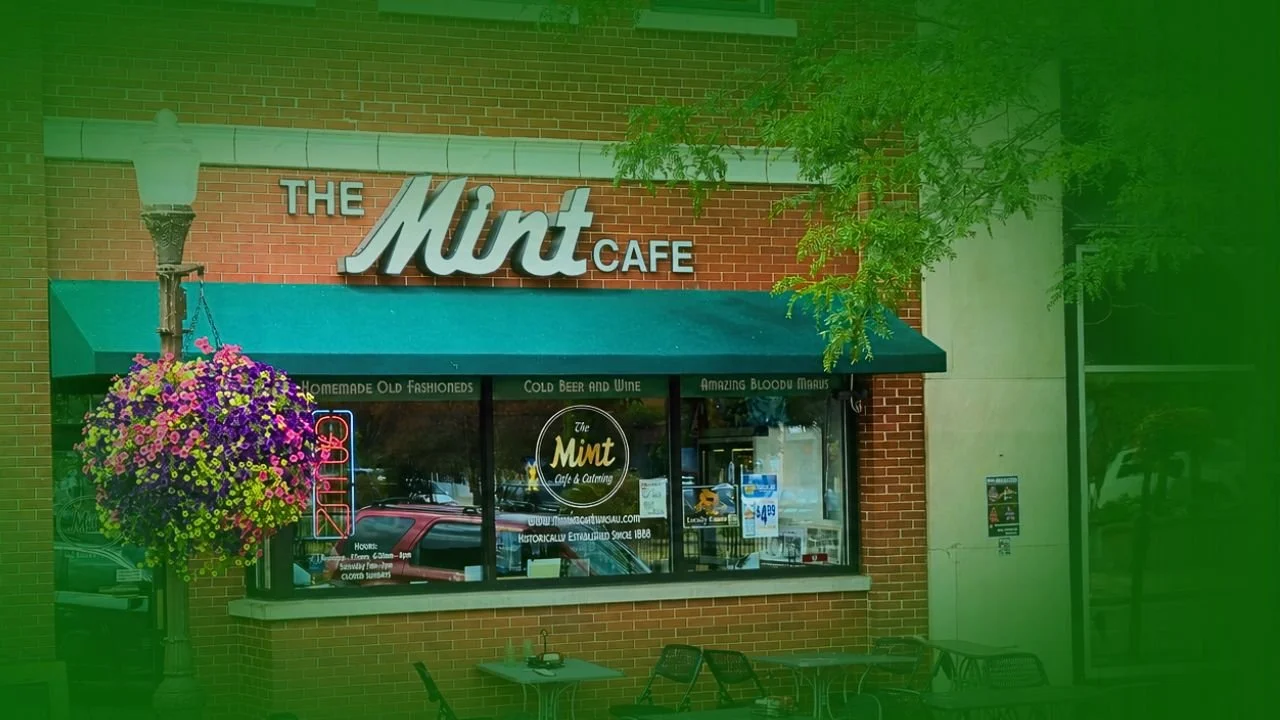 Front of The Mint Cafe with brick exterior, green awning, hanging flower basket, sidewalk tables, and a neon sign in the window.