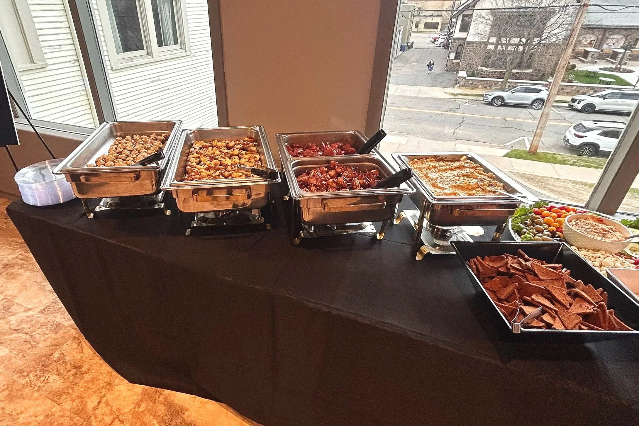 Buffet table with various dishes, including meats, vegetables, and other side dishes, set near a large window overlooking a street with parked cars.