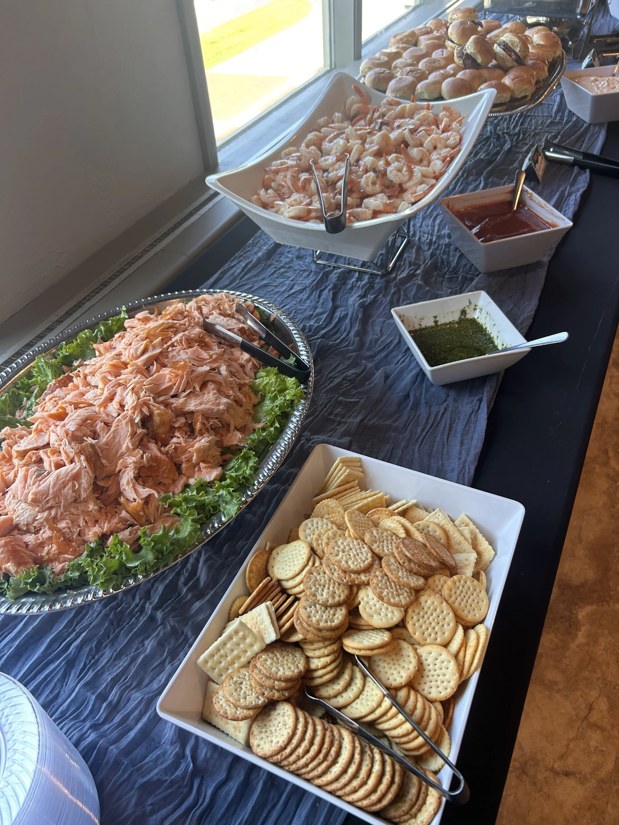 Buffet table with trays of crackers, shrimp, chicken, slider buns, and condiments near a window.