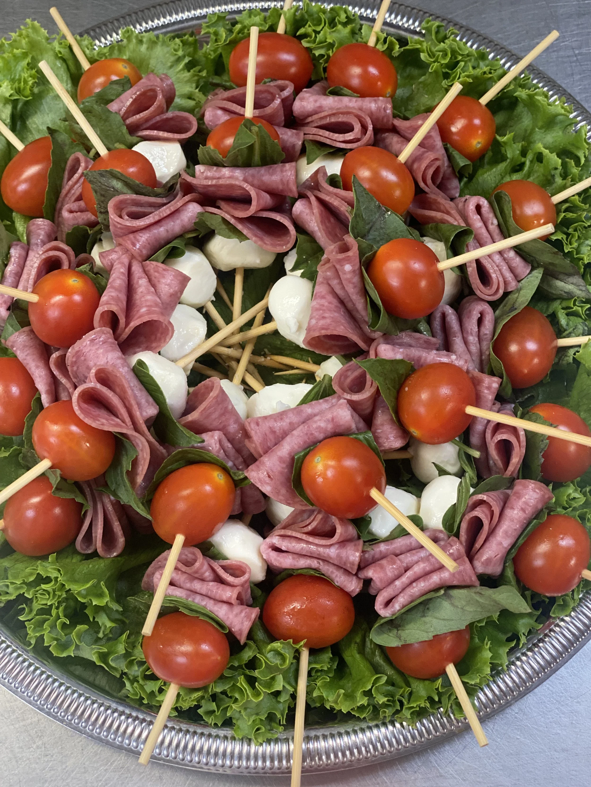 A silver tray holding skewers of cherry tomatoes, mozzarella balls, slices of salami, and basil leaves, with green lettuce and curly lettuce around the edges.