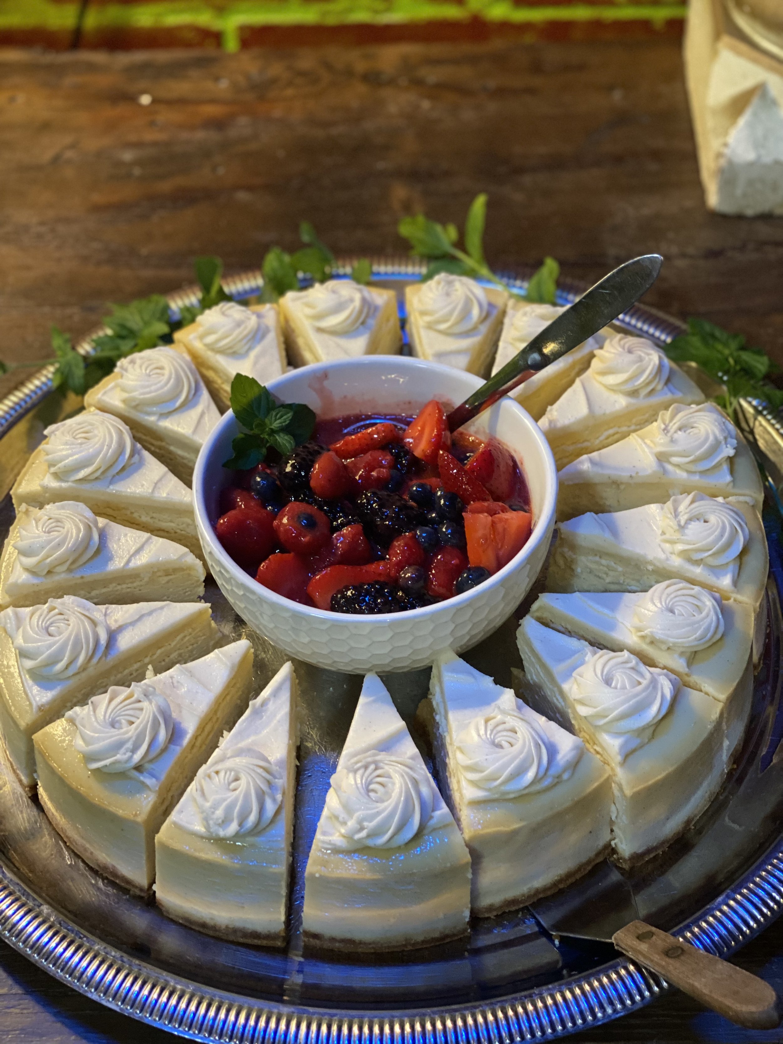 A round tray of sliced cheesecake with spiral decorations on top, topped with a bowl of mixed berries and sliced strawberries in the center.