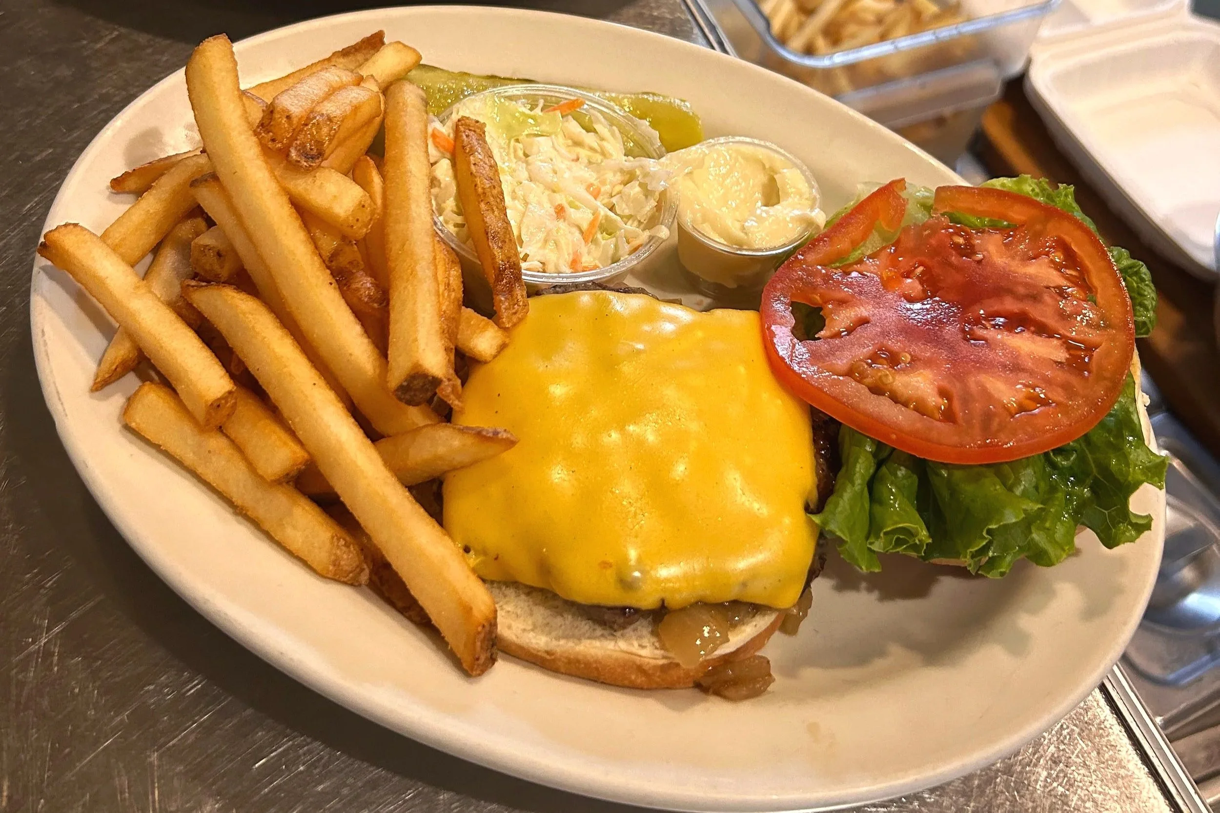 A plate of food with French fries, a cheeseburger topped with cheddar cheese, lettuce, tomato, and pickles, along with side condiments including mayonnaise, coleslaw, and mustard.