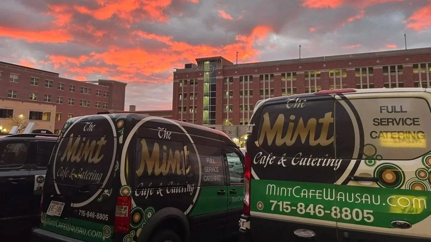 Two delivery vans with branding for The Mint Cafe & Catering parked outside a large red-brick building at sunset, with a colorful orange and pink sky.