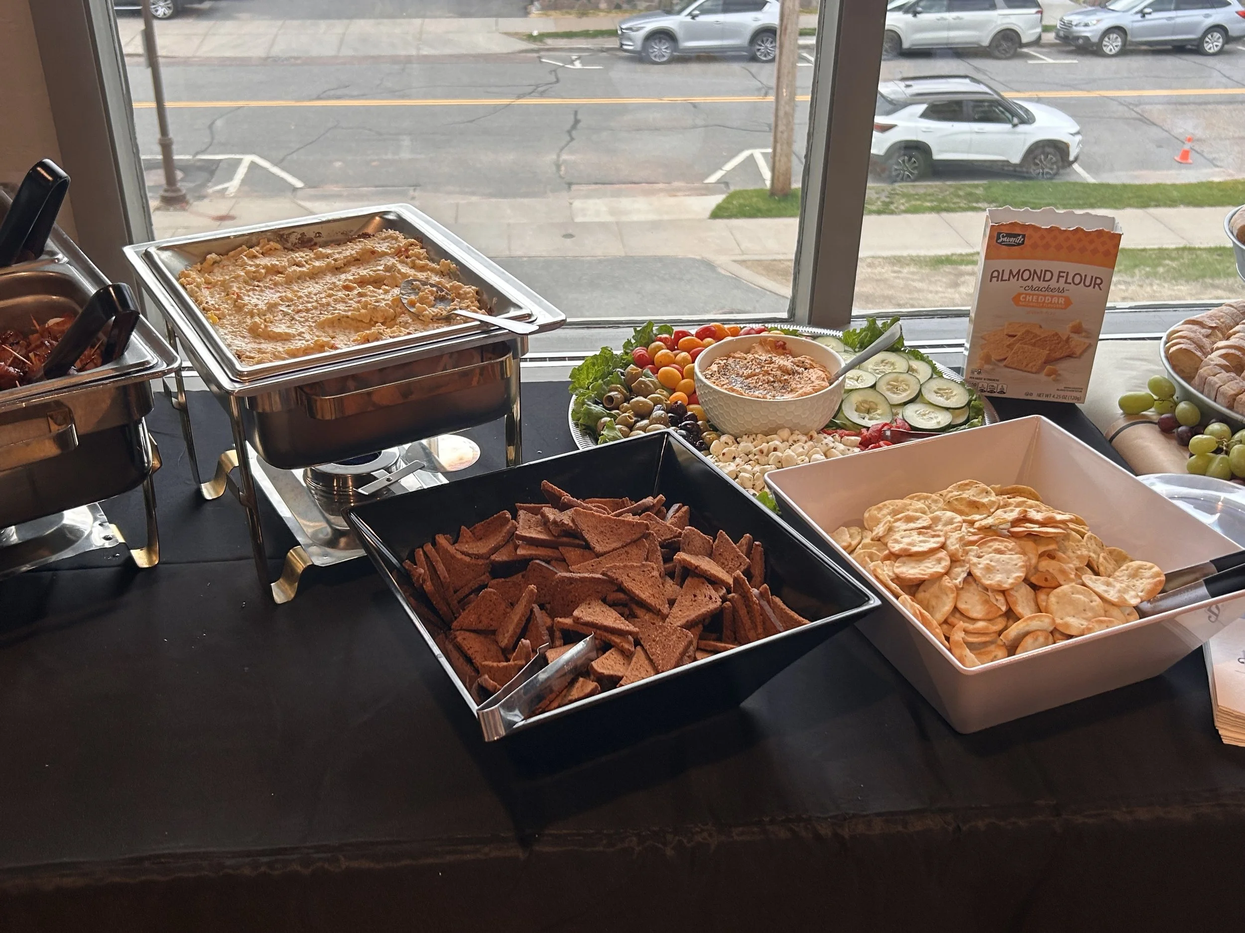 A buffet table with various food items including crackers, vegetables, dip, and bread, set against a large window overlooking a street with parked cars and a traffic cone.