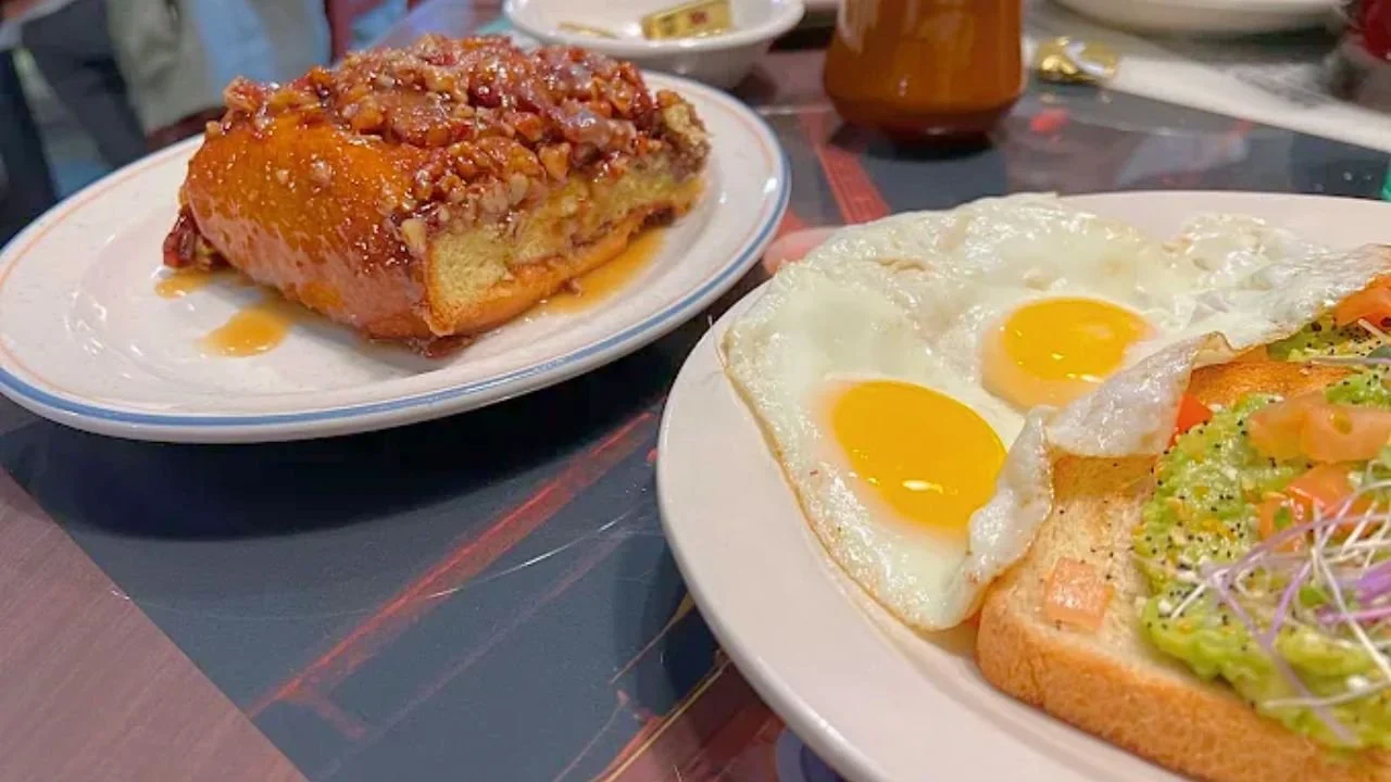 Slice of cherry coffee cake with crumb topping on a white plate and plate with two fried eggs and toast with avocado, tomato, and sprouts on a table.