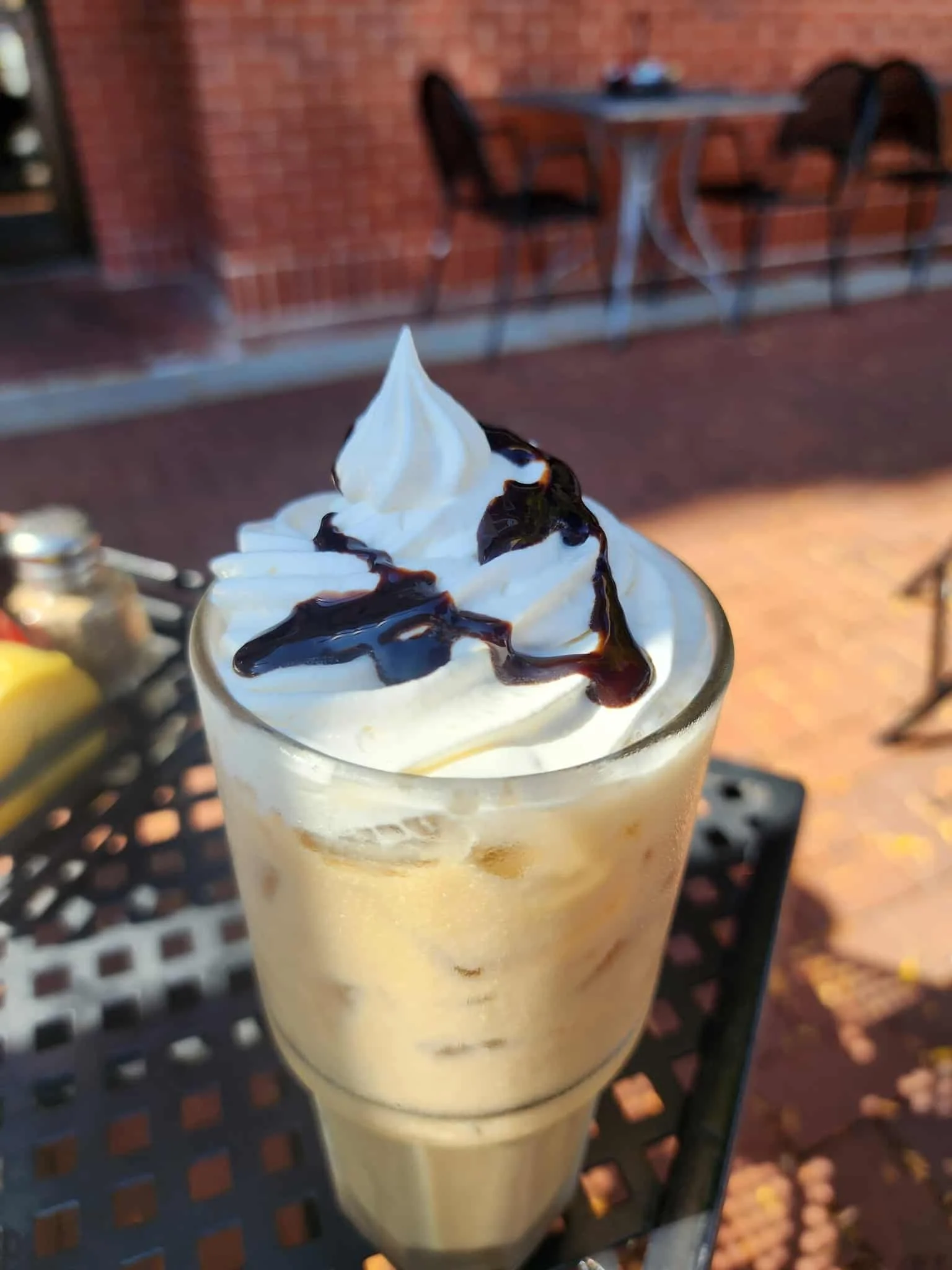 A glass of vanilla milkshake topped with whipped cream and chocolate syrup, sitting on an outdoor table.