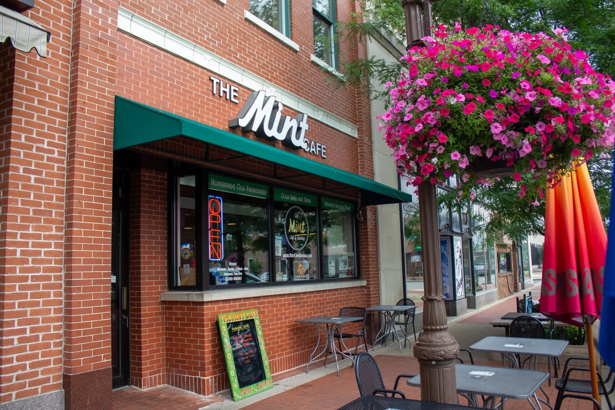 Outside view of a brick café named 'The Mint' with a green awning, pink flowers hanging from a streetlamp, sidewalk tables and chairs, and a chalkboard sign.