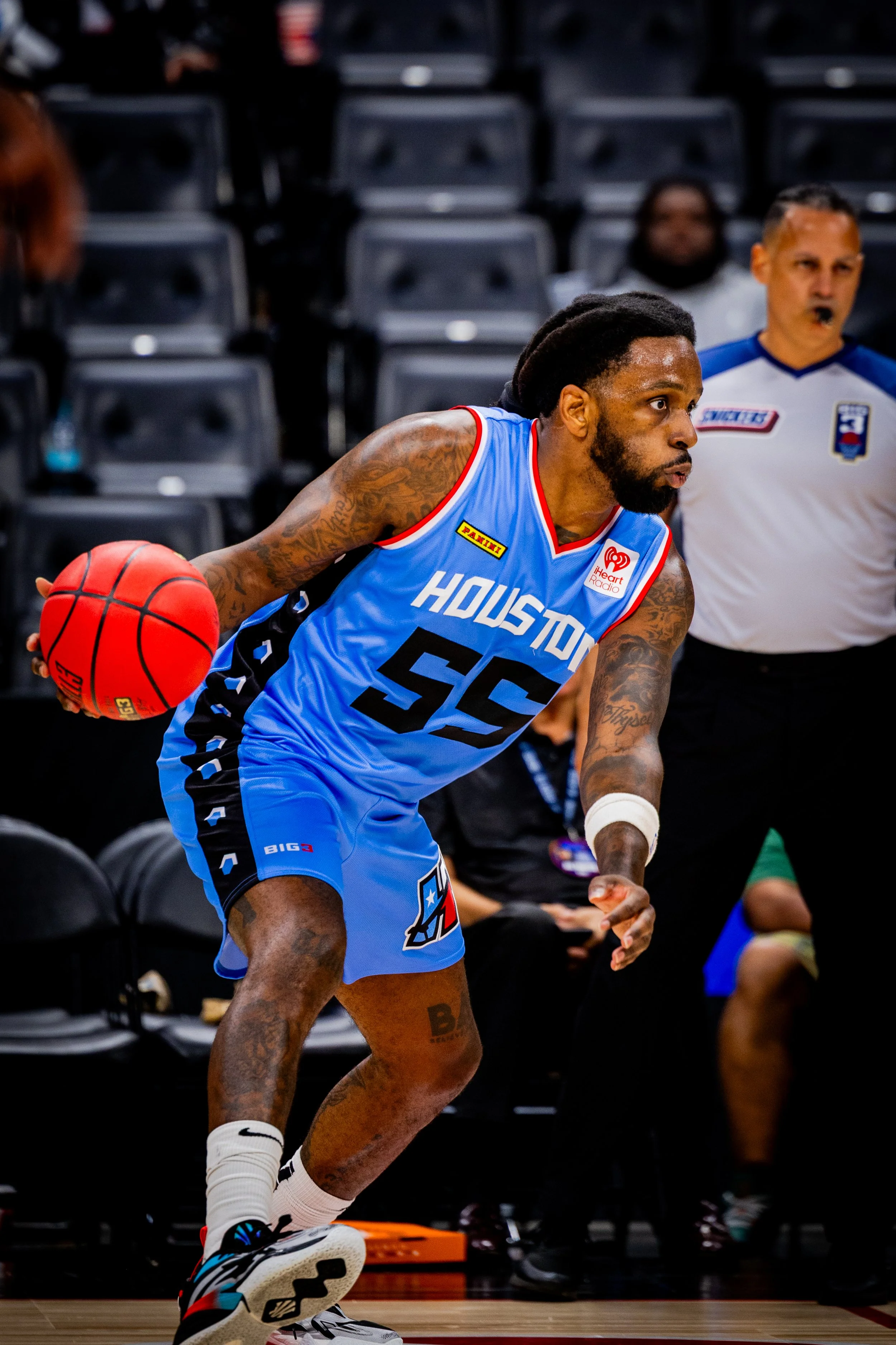 A male basketball player in a blue Houston Righands jersey dribbling a basketball during a game with an official and spectators in the background.