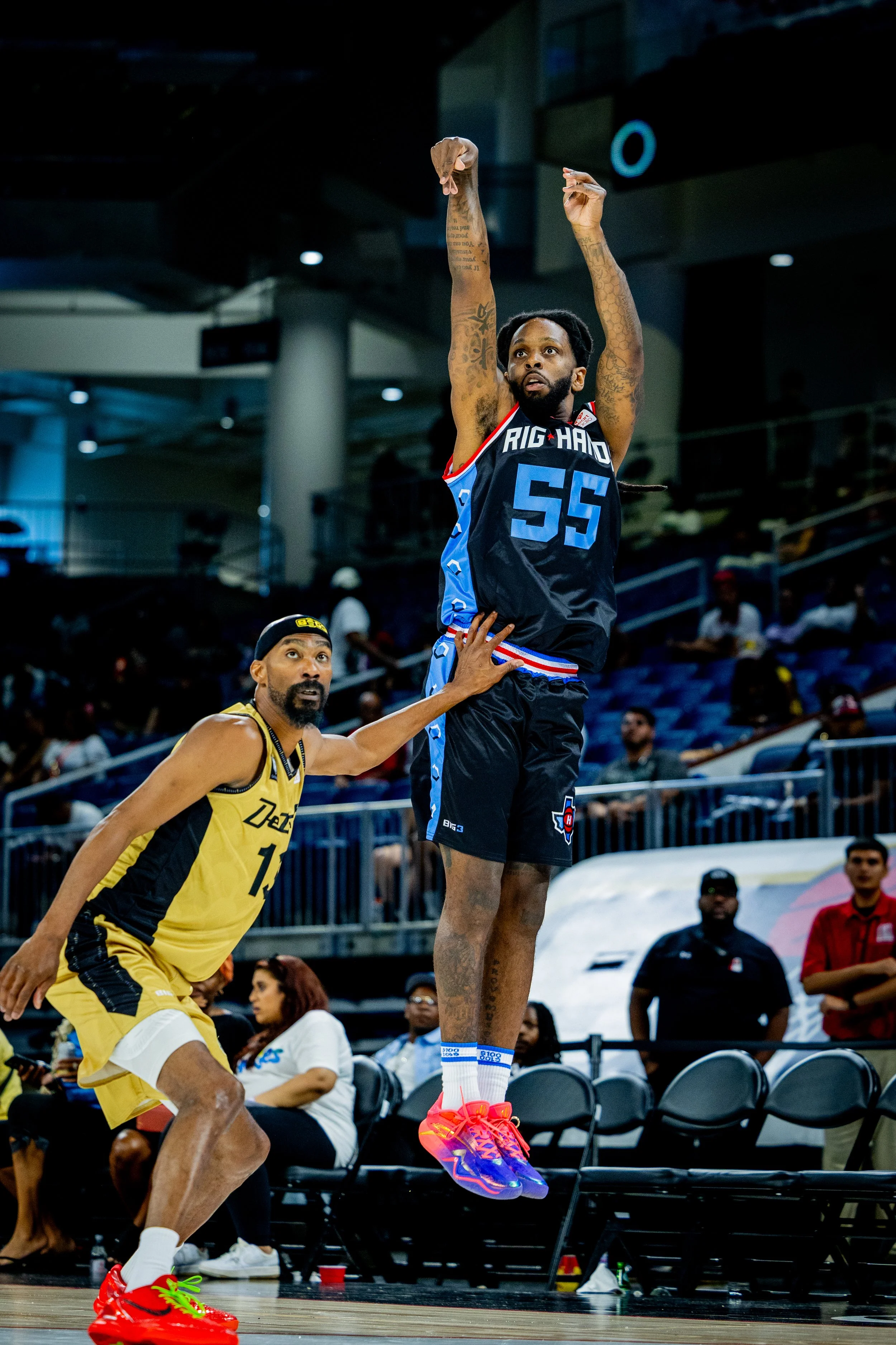 Basketball player in a black and blue uniform jumping to make a shot during a game while an opponent in a yellow uniform guards him near the basket.