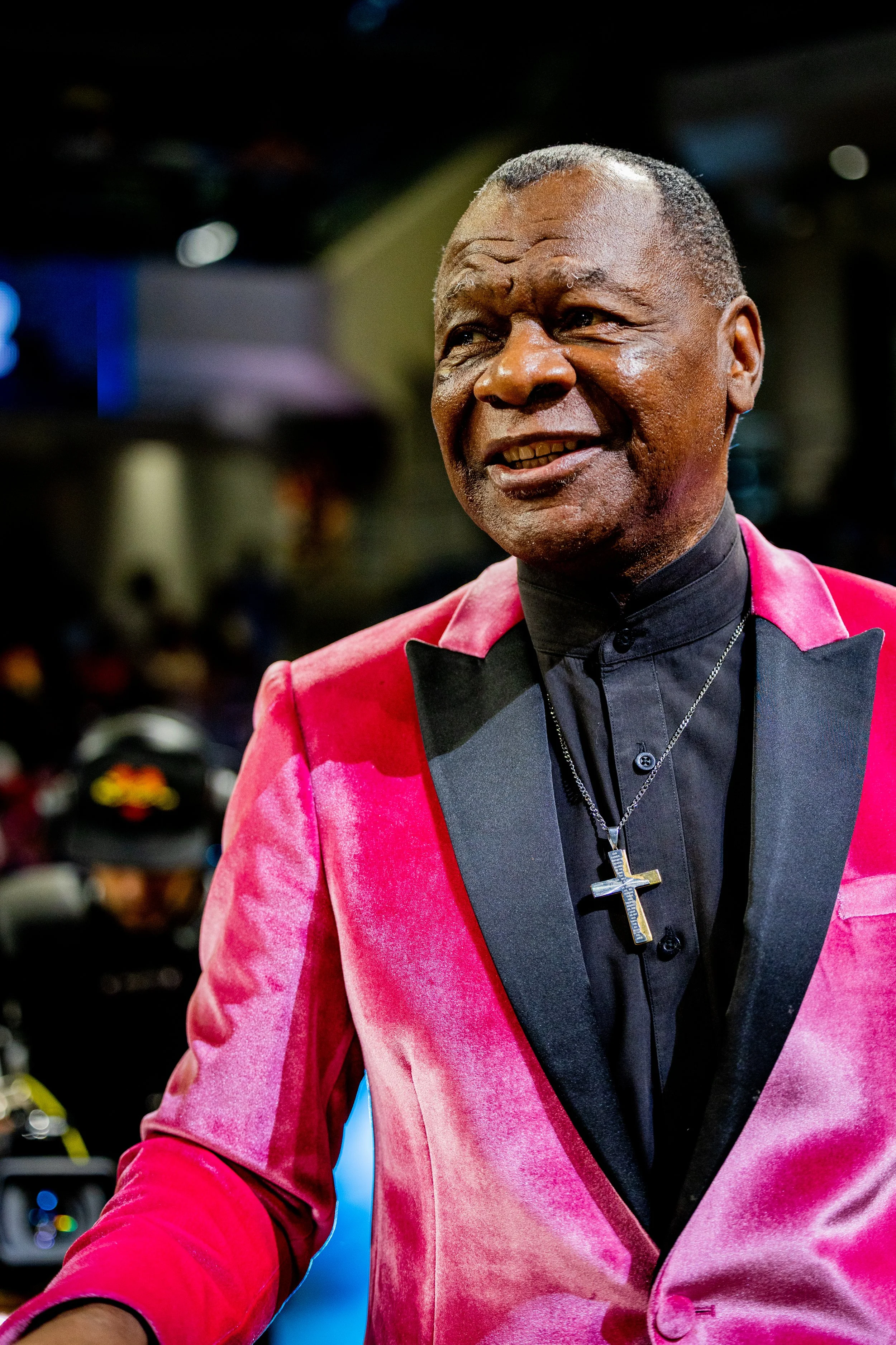 A middle-aged man with a warm smile, wearing a black shirt with a collar, a pink velvet blazer, and a silver cross necklace. The background is blurred with lights and possibly a crowd, suggesting he is at an event or gathering.