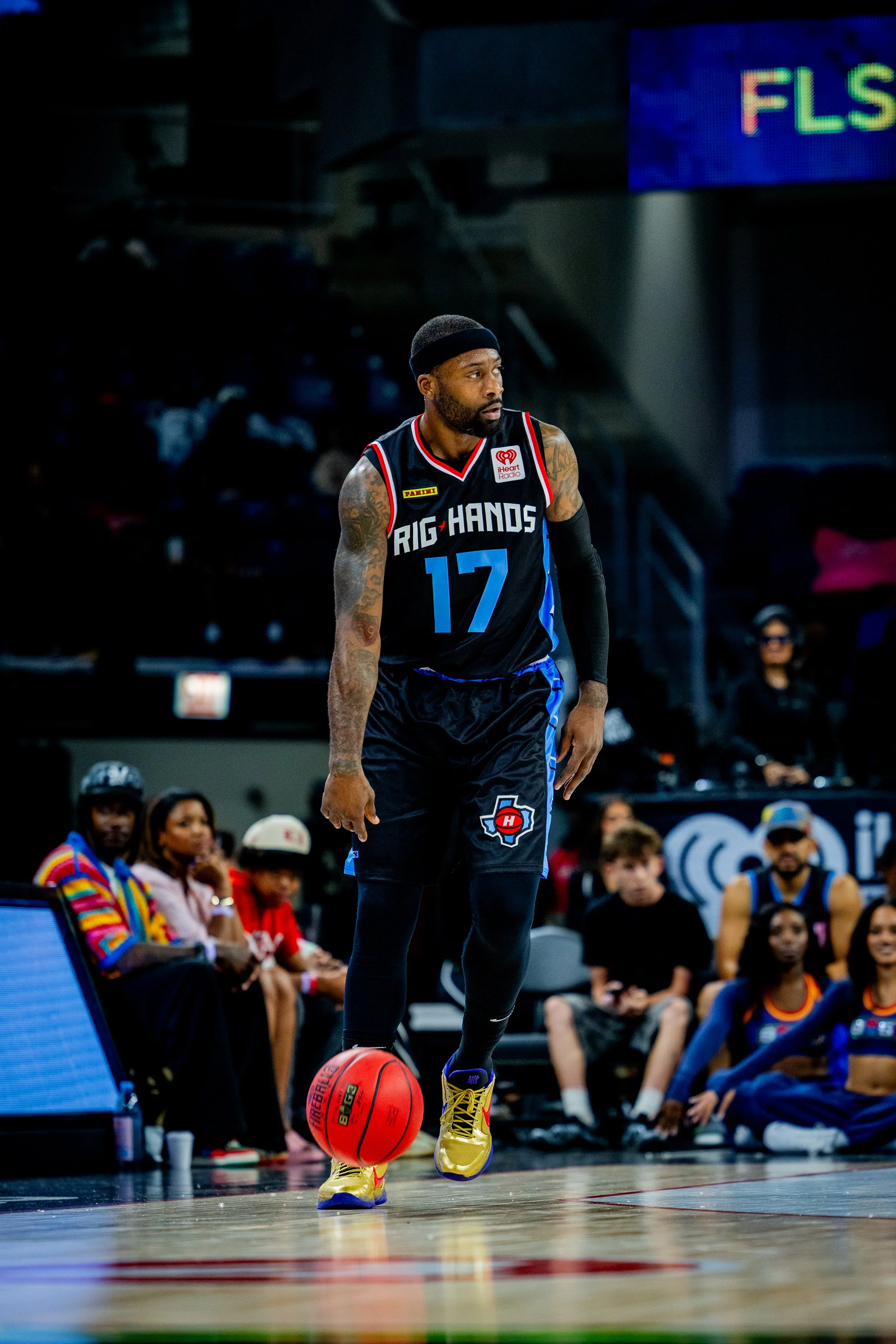 A basketball player wearing a black jersey with 'RIG HANDS' and the number 17 is dribbling a bright orange basketball on a court, with spectators and other players seated in the background.