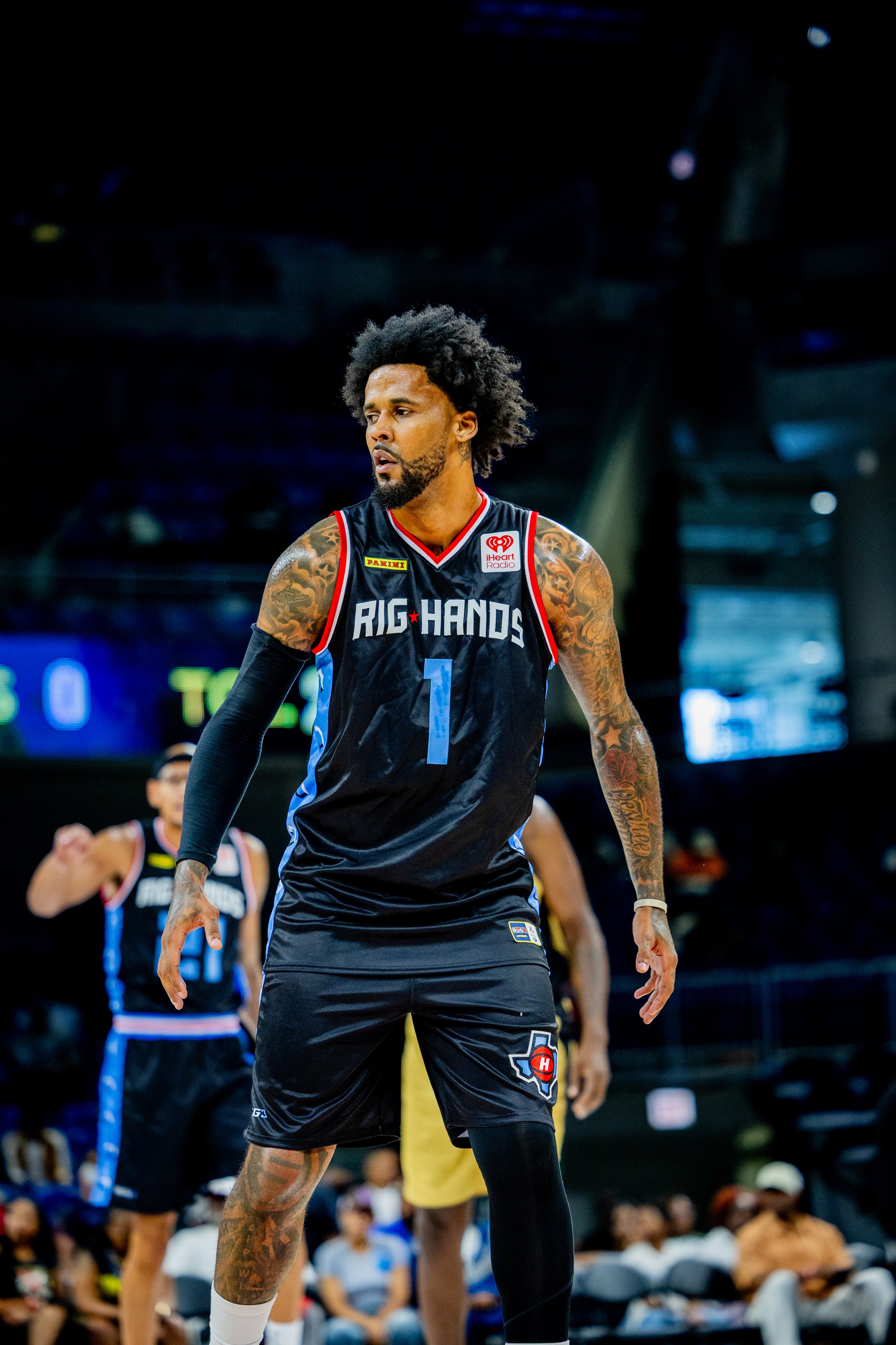 A basketball player with tattoos, wearing a black jersey with blue, red, and white accents, stands on the court during a game. He has an afro hairstyle and a beard, with a focused expression.