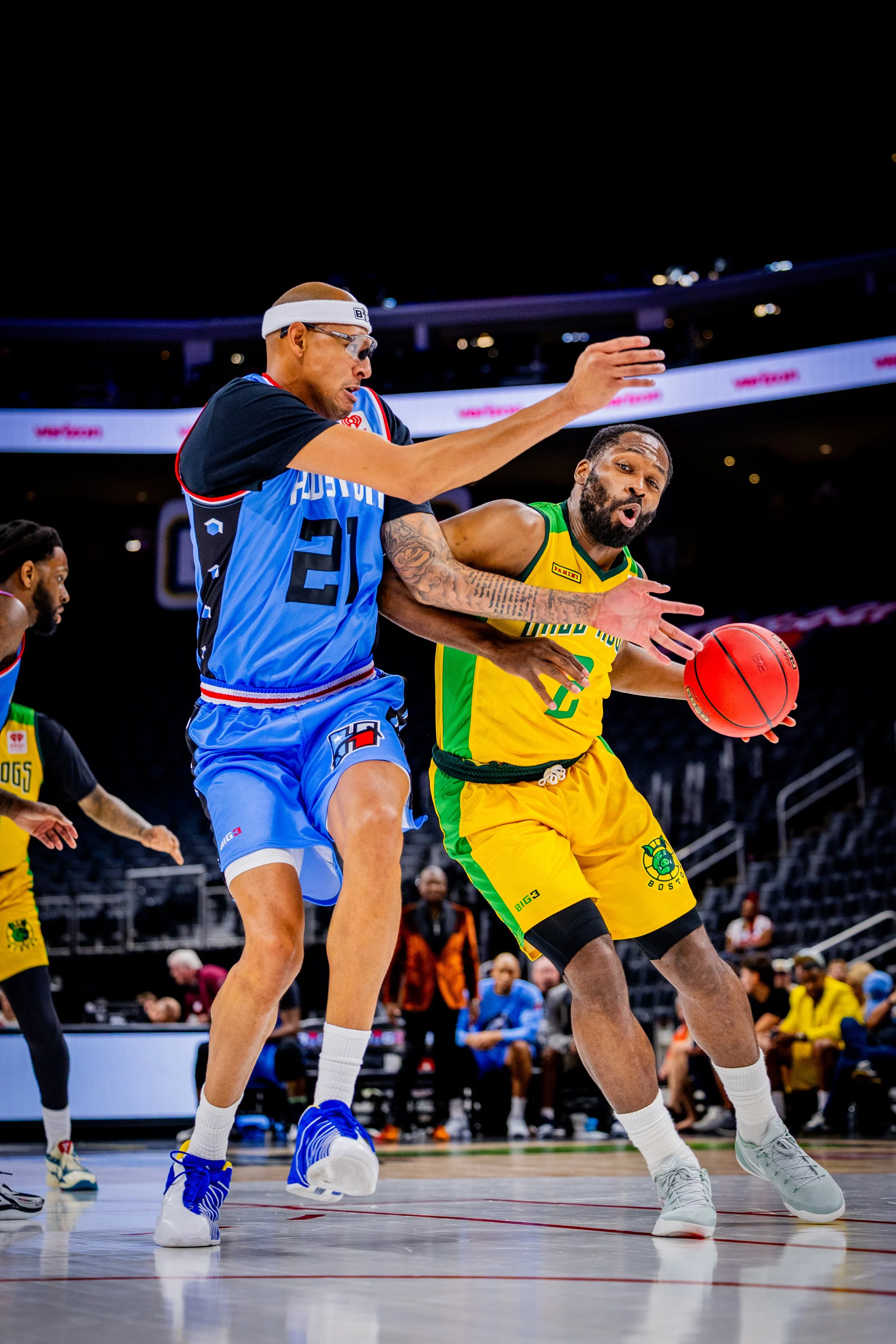 Two basketball players are battling for possession of the ball during a game, with others and spectators in the background.