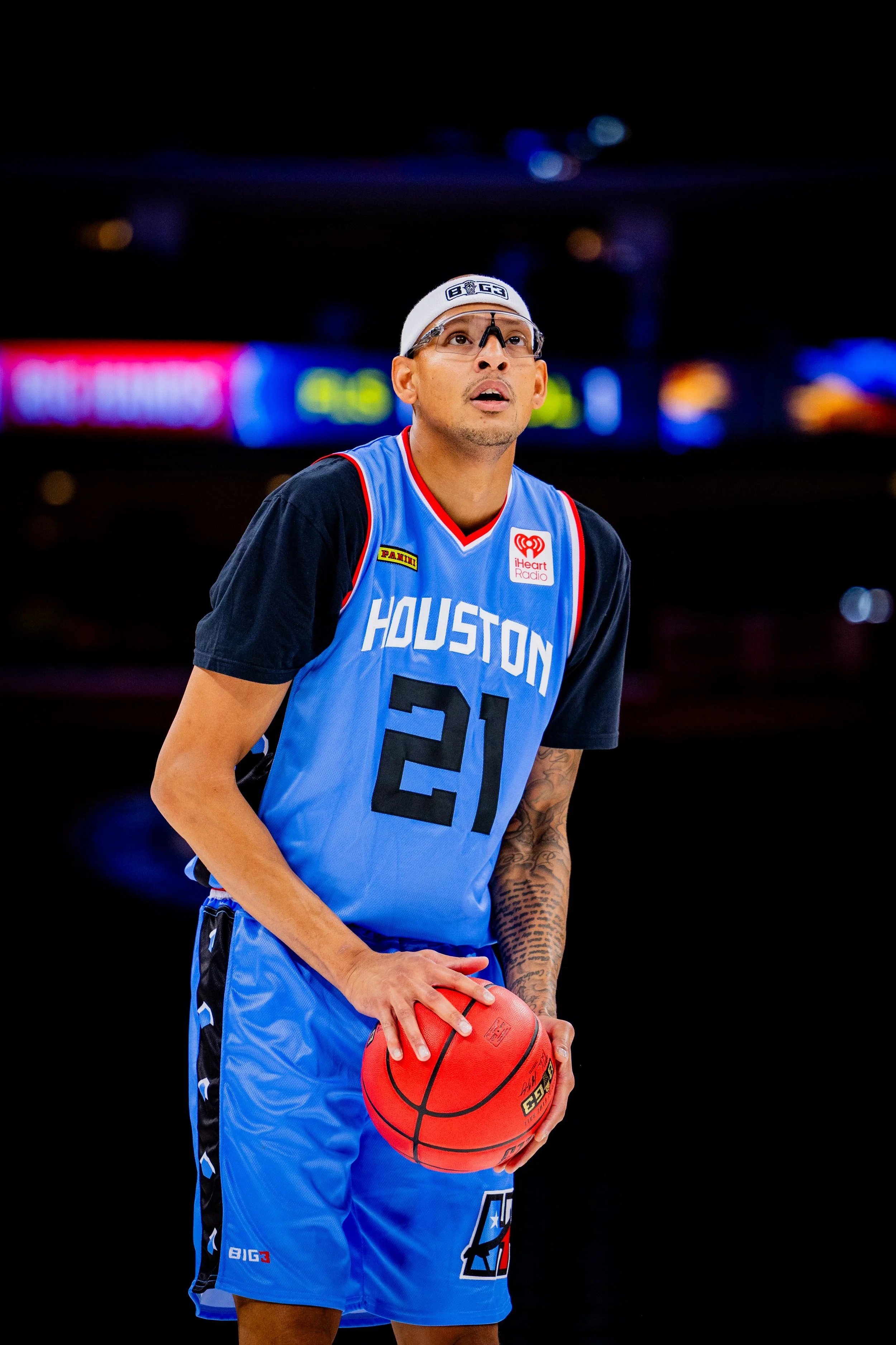 A male basketball player in a blue Houston jersey with the number 21, holding a basketball, on a basketball court at night with bright digital advertisements in the background.