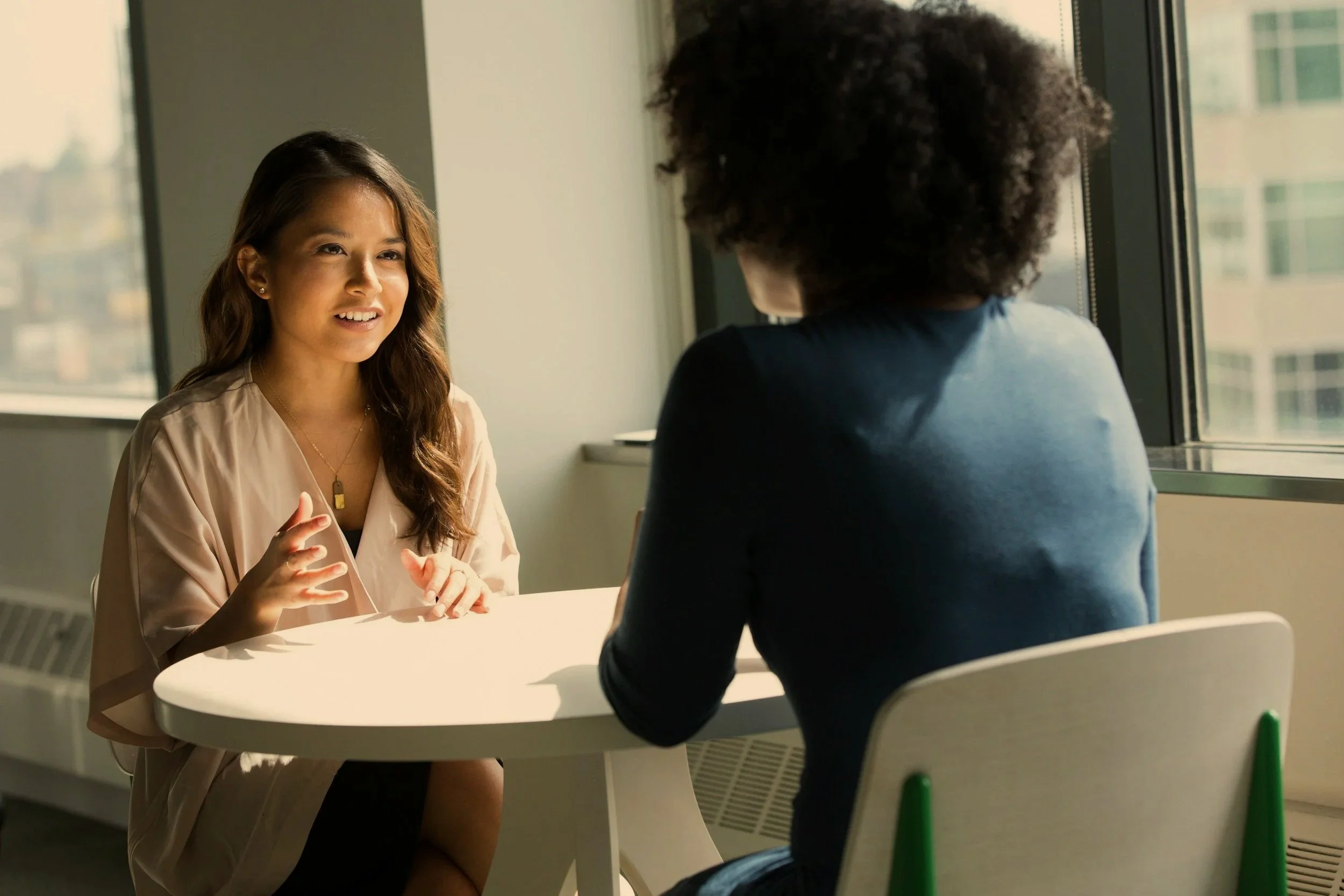 Two women are seated at a small table near a window having a conversation. One woman, facing the camera, has long brown hair, is wearing a beige wrap dress, and is gesturing with her hands. The other woman has curly hair, is wearing a dark top, and has her back to the camera. The setting appears to be a modern office or lounge area with natural light coming in from the window.