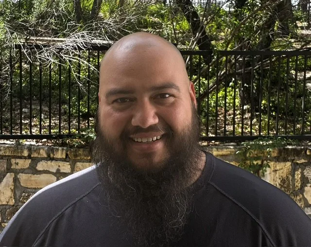 A man with a bald head and full beard smiling outdoors, with a black metal fence, stone wall, and trees in the background.