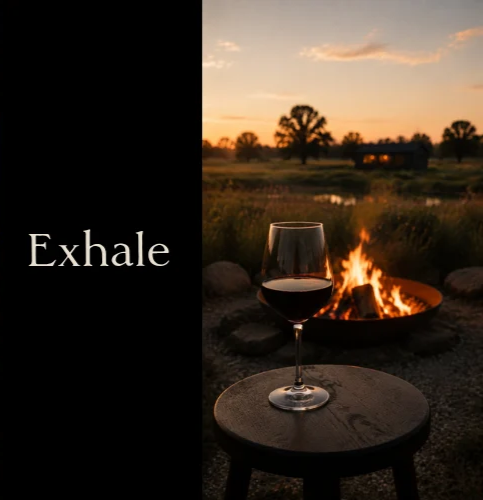 A glass of red wine on a small round table outdoors at sunset, with a campfire and a rural landscape in the background.