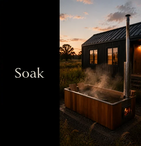 A hot tub outdoors at sunset with steam rising from the water, next to a black house with metal roof and a chimney, surrounded by grassy landscape and trees.