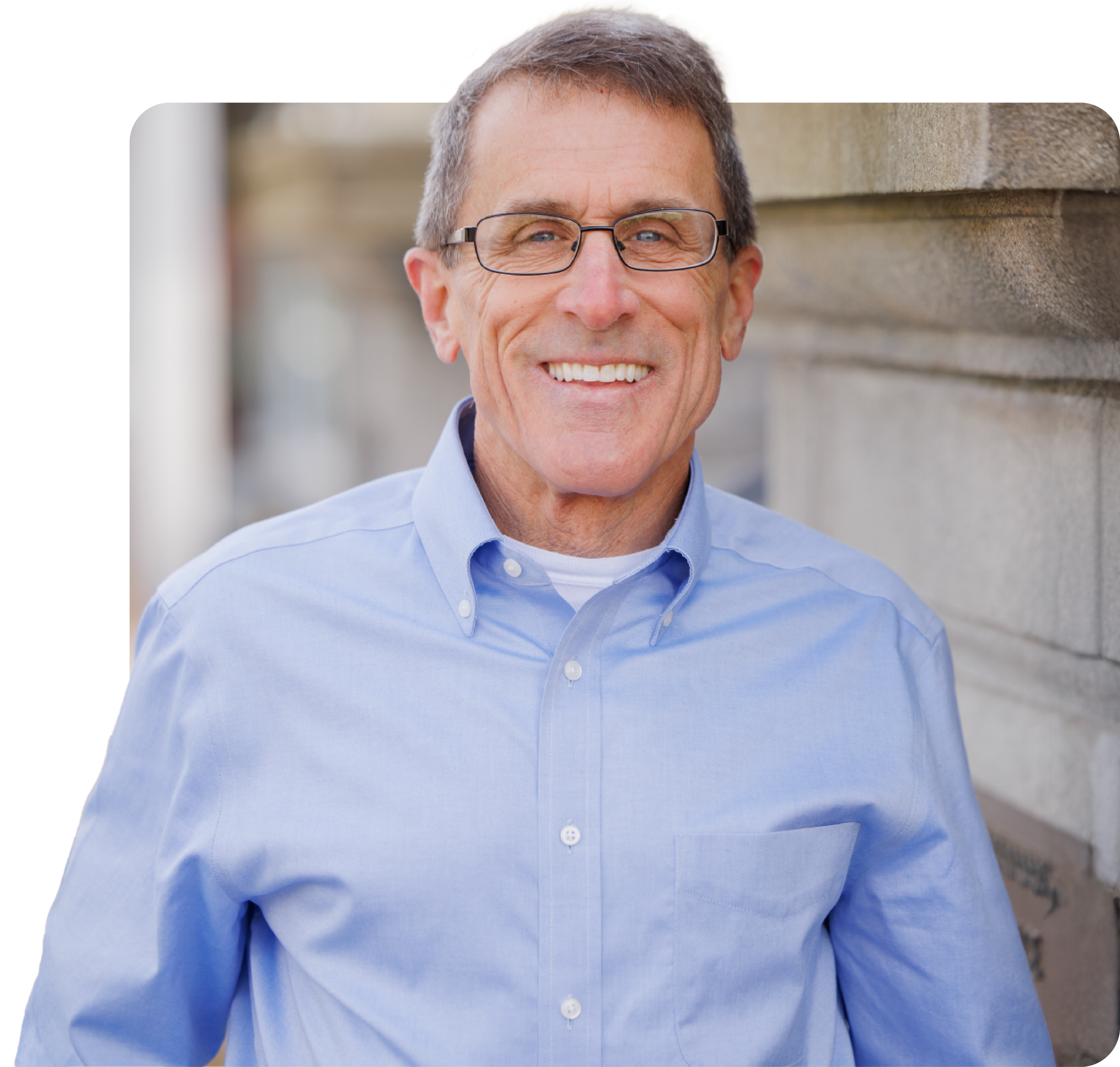 Close-up of a smiling middle-aged man with gray hair wearing glasses and a light blue button-down shirt, standing outdoors next to a concrete wall.