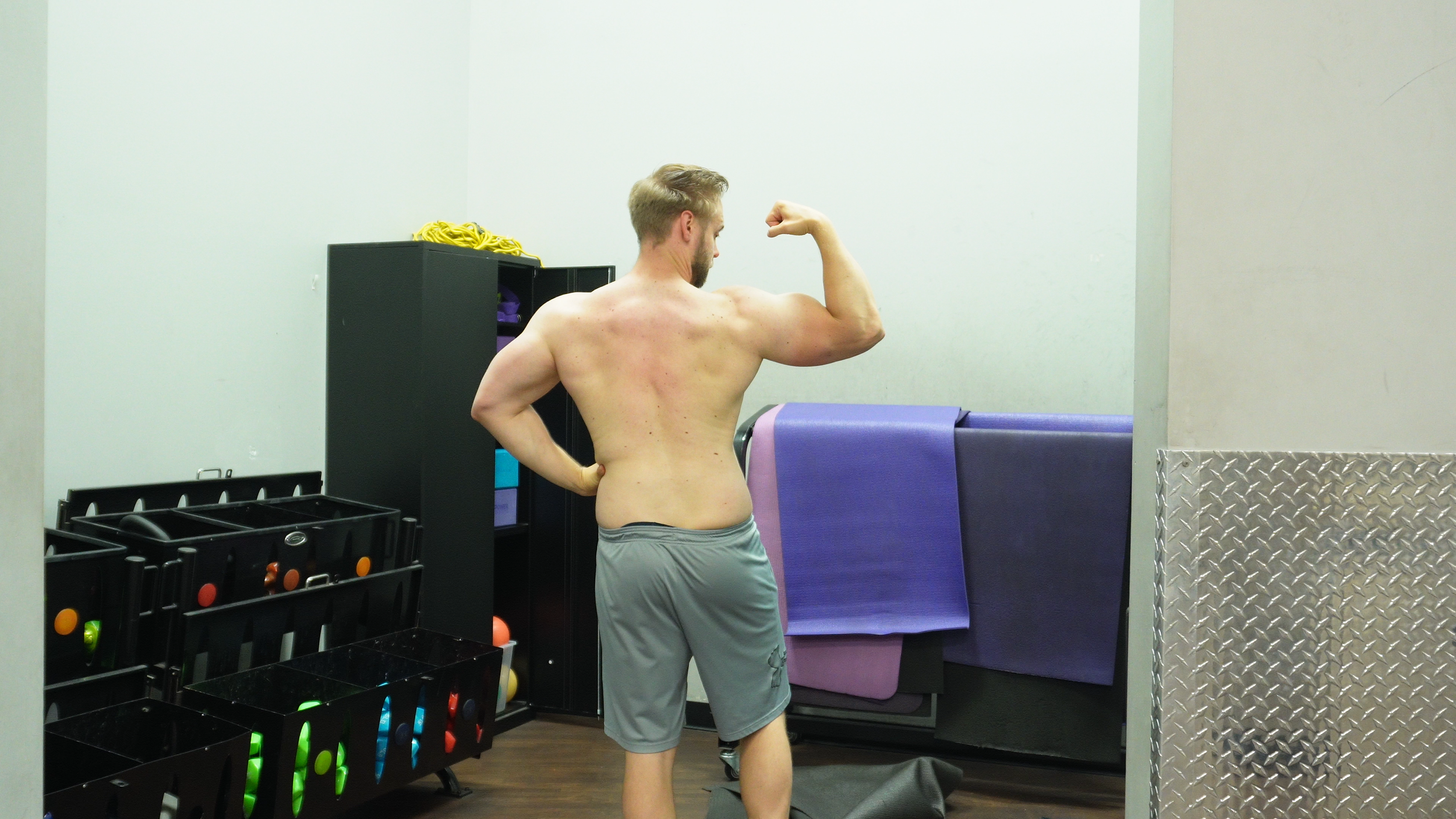 A muscular man flexing his right arm in a gym, facing away from the camera, with gym equipment and mats in the background.