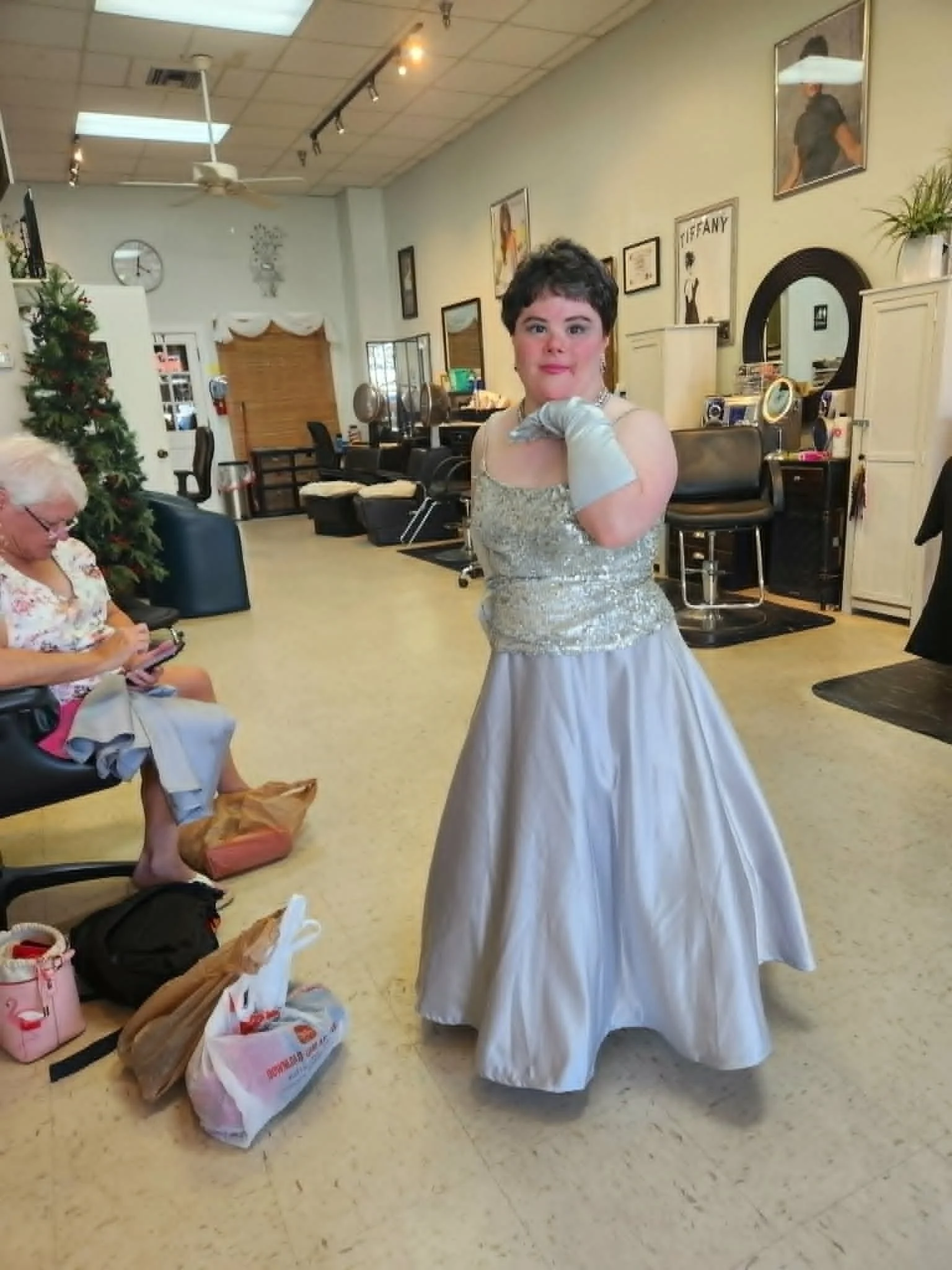 A woman in a fancy silver and white gown posing indoors in front of a mirror. She is wearing long gloves and a necklace. There are people sitting and bags on the floor nearby, and the background features framed pictures and a decorated Christmas tree.