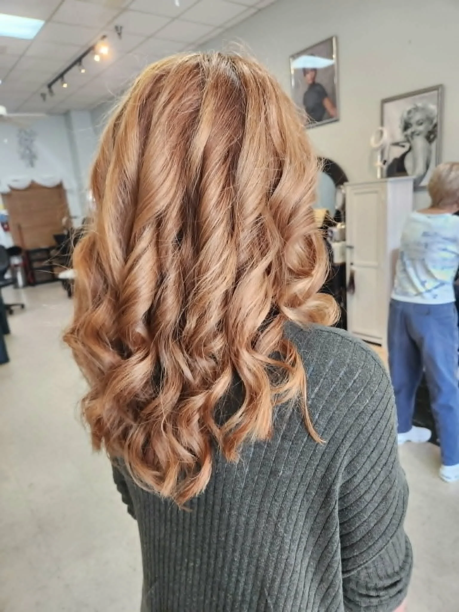 Back of a woman with shoulder-length, wavy, blonde hair, wearing a gray knitted sweater, in a hair salon with framed photos on the wall and other people in the background.