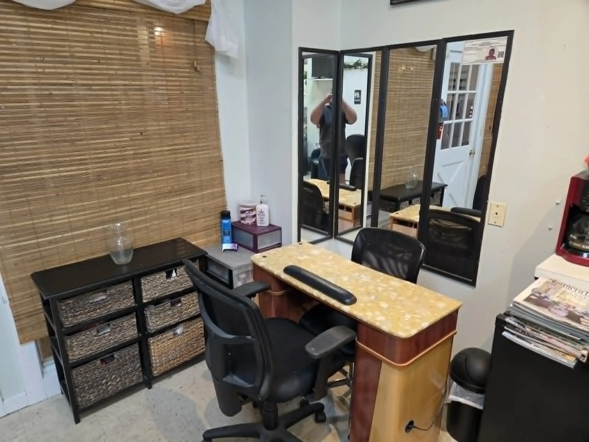 Interior view of an office or reception area with a desk and chairs, a wall with a bamboo style covering, a mirror, and various items including a water bottle, hand sanitizer, and stacks of papers.