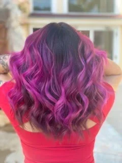 Woman with wavy hair dyed in shades of purple and pink, wearing a red top, facing away outdoors.